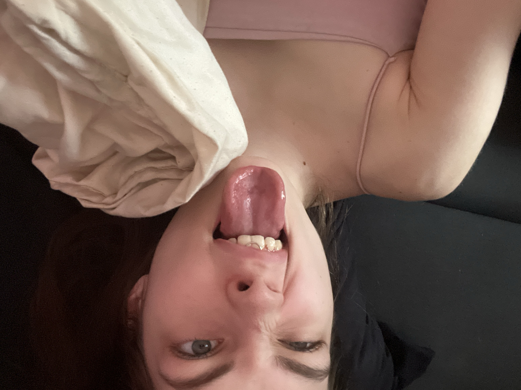 A woman is lying upside down with her tongue sticking out and one eye squinting. She is holding a white blanket and wearing a pink tank top. She appears to be on a black surface, possibly a bed or couch. The location is not identifiable as Saint Petersburg, Russia based on the visual information provided.