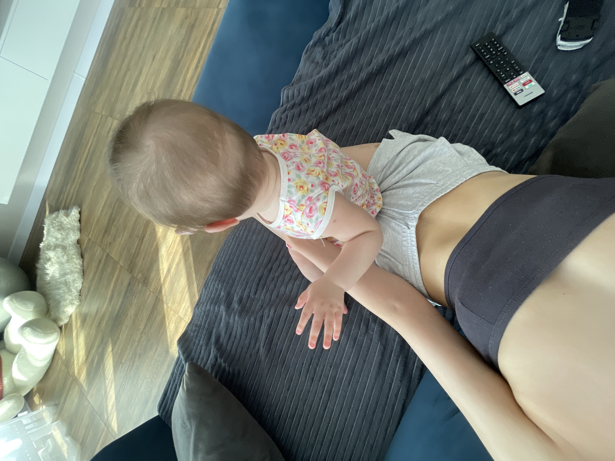 The image captures an overhead view of a baby interacting with an adult in an indoor setting in Podshivalovo, Russia. An adult, seen from the torso, is lying down, wearing a grey t-shirt and a black bra or sports top. A baby, dressed in a white floral sleeveless top and grey shorts, is positioned above the adult, with their hands outstretched towards a dark grey textured blanket covering a blue sofa or bed. The baby's light brown hair is visible from the top of their head.
The setting includes a wooden floor where bright sunlight streams in, casting patterns of light and shadow, indicating daytime. Visible objects on the floor include a white llama-like stuffed toy, another white soft toy, and a transparent plastic container. On the dark grey blanket, a black remote control is visible, featuring various buttons such as "MENU," "VOL," "CH," "OK," and "POWER." A pair of white socks is also on the blanket. A grey throw pillow is seen to the left. The scene depicts a relaxed domestic moment.