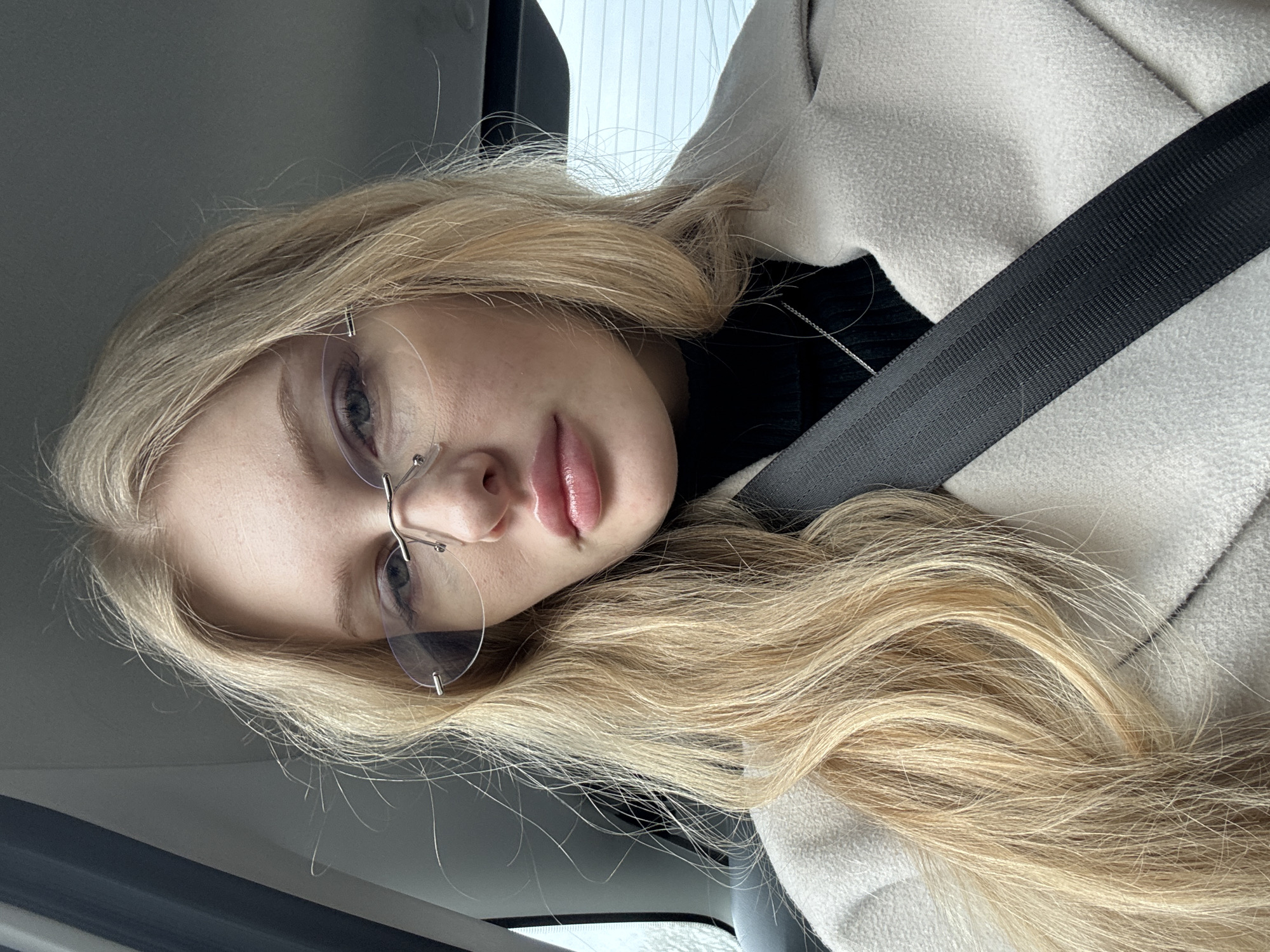 A fair-skinned woman with blonde hair is shown inside a vehicle. She is wearing rimless glasses, a black turtleneck sweater, and a light-colored coat. A seatbelt is visible across her body. The location is unspecified and there's no direct evidence for the city of Perm, Russia.