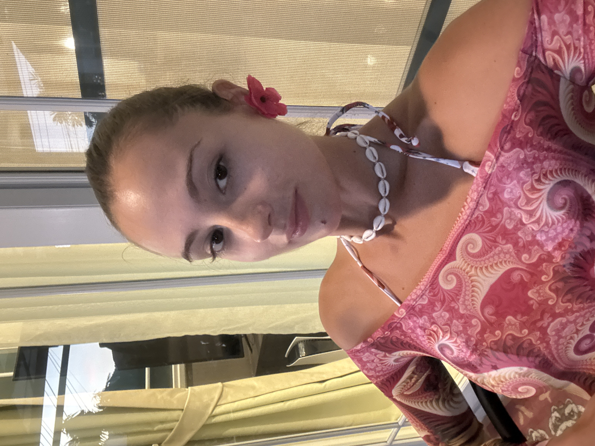 A young woman poses indoors in Chalong, Thailand. She is wearing a pink patterned top and a seashell necklace. A red flower adorns her hair. The background features blinds and a partial view of the interior.
