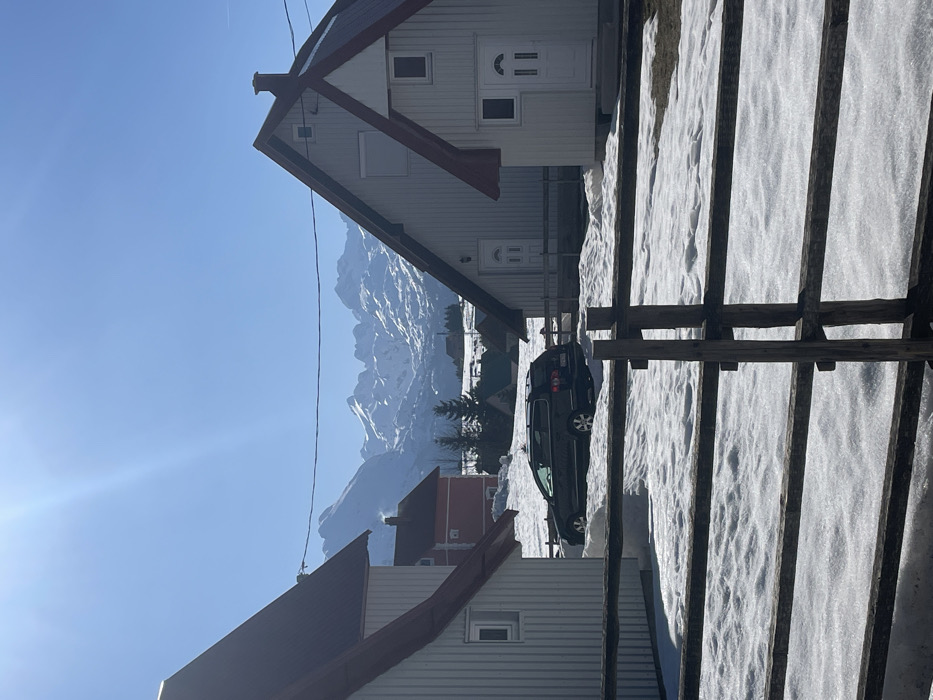 The image depicts a winter or early spring scene in Palež, Montenegro, under a clear, bright blue sky with direct sunlight. In the immediate foreground, a weathered, dark brown split-rail wooden fence with multiple horizontal rails extends across the lower portion of the frame. Beyond the fence, the ground is covered in uneven patches of melting snow, revealing some bare earth.
The midground features three distinct houses with white vertical siding and steep reddish-brown gable roofs. On the far left, a two-story house has one visible window on its left side. To the right, a larger two-story house exhibits two white doors on its front facade, suggesting multiple entrances, and a single small window above the right door. A chimney is visible on its roof. Partially obscured on the far right is a third, smaller white house with a similar roof style and one visible white door.
A dark-colored, possibly black, station wagon is parked between the left and center-right houses, partially embedded in the snow, facing away from the viewer. Further in the midground, a red-colored house is visible between the foreground structures, nestled among snow-covered terrain and sparse evergreen trees. A thin black utility line is visible stretching horizontally across the upper midground, partially obscuring the distant mountains.
The background is dominated by a majestic range of rugged, snow-capped mountains under the clear sky. The mountains exhibit prominent white snow coverage interspersed with darker rock formations. The sun's glare is visible as a bright lens flare in the upper left quadrant of the sky. No individuals or animals are present in the scene, and no specific actions or interactions are depicted.