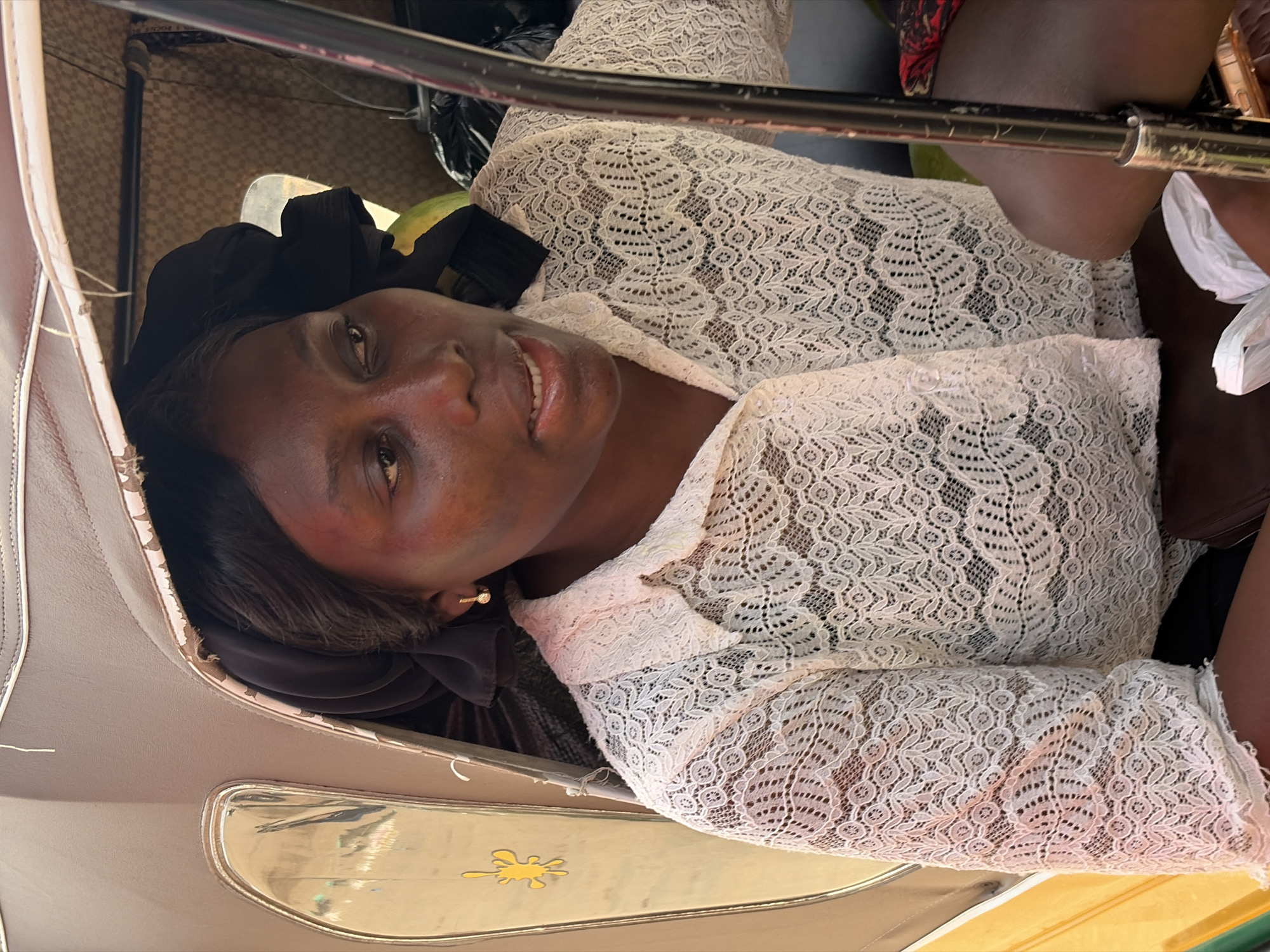 In Gura Top, Nigeria, a woman is seated inside the cabin of a motorized tricycle, also known as an "okada" or "boda-boda". She is looking to her right and speaking. She wears a white lace blouse and a black headscarf. Behind her, several black plastic bags are visible within the tricycle's cabin. A chrome-colored bar runs vertically in the foreground, partially obscuring her right side. The exterior of the tricycle, visible on the left, is yellow and brown with a reflective silver trim. A yellow splat-shaped sticker is affixed to the exterior. The interior of the tricycle is lined with brown material.