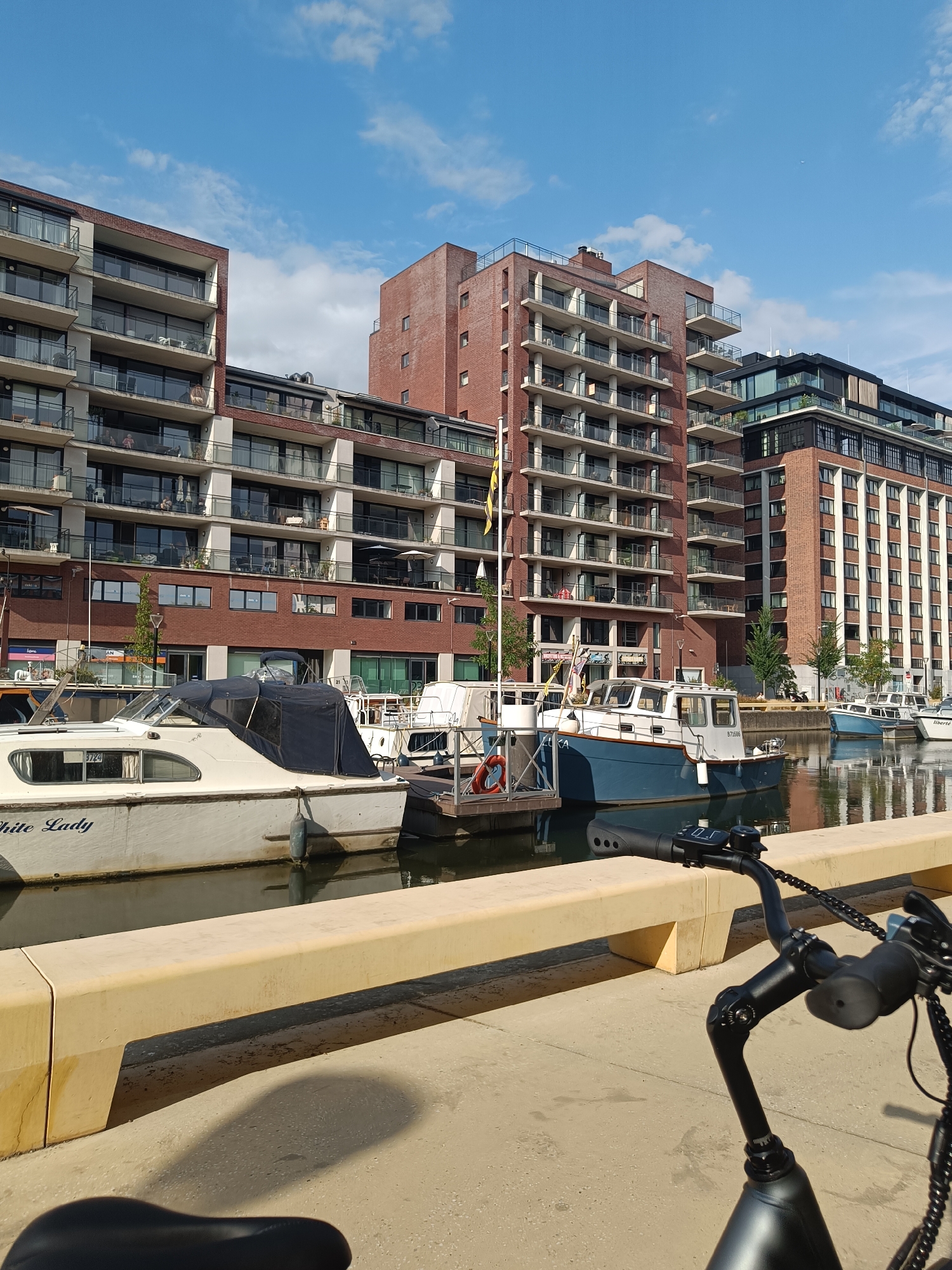 The image depicts a waterside view of modern apartment buildings and docked boats on a sunny day. The buildings are multi-story structures with a predominantly brick facade and numerous balconies with glass railings. The boats are of various sizes, with one prominently featuring the name "White Lady" on its hull. The water is calm and reflects the buildings and sky. In the foreground, a portion of a bicycle is visible, suggesting it is the viewpoint from which the photo was taken. The overall setting appears to be a well-developed urban area with a marina or waterfront district. This scene is located in the city of Leuven, Belgium.