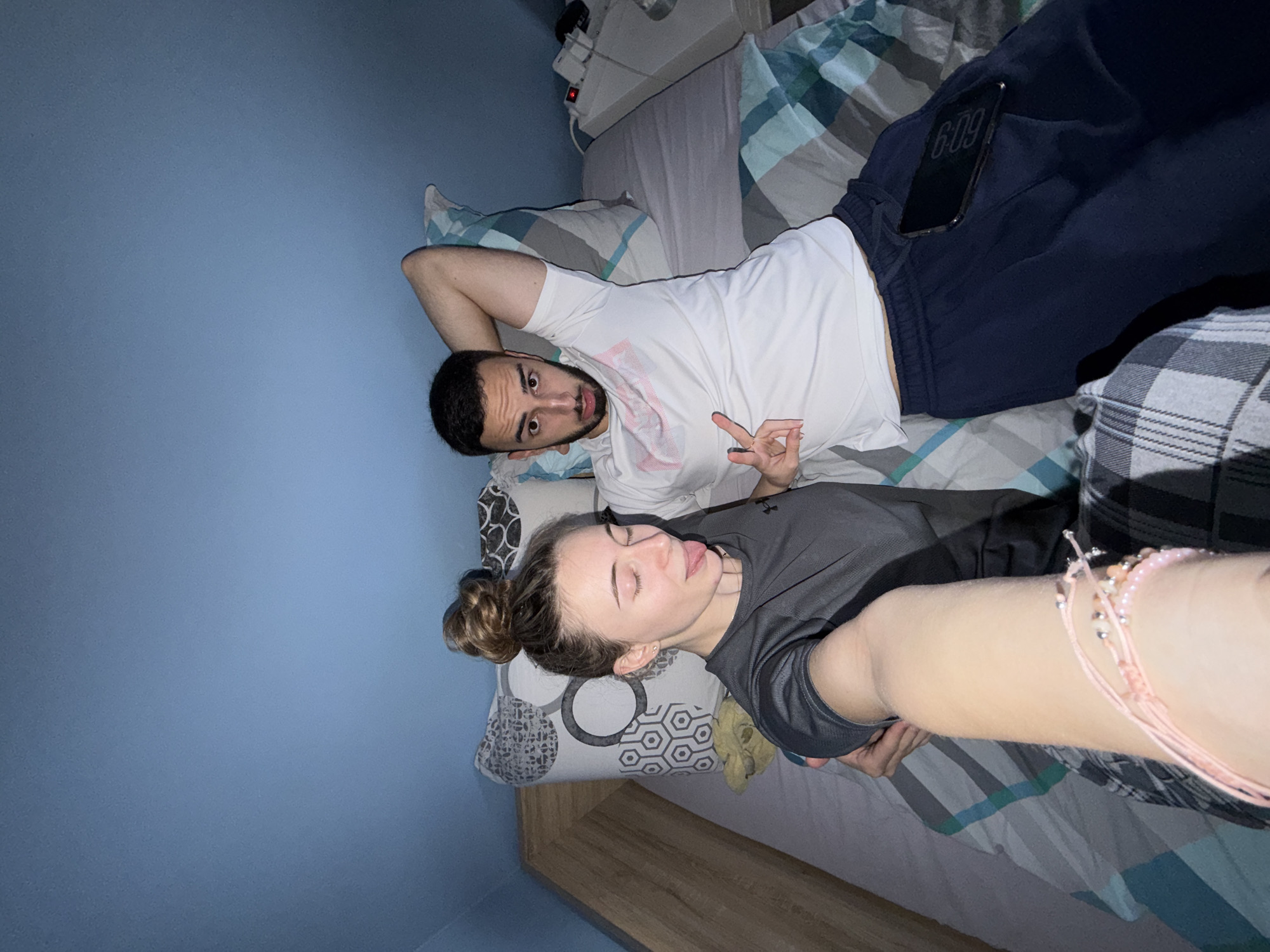 Two individuals lie on a bed with patterned bedding, possibly indoors in a residence. The man wears a white t-shirt and dark pants. He is reclining and making a peace sign. A smartphone is visible in his pants pocket, displaying the time as 6:09. The woman, closer to the camera, wears a dark-colored shirt. Her hair is in a bun. Her arm extends towards the camera, adorned with pink bracelets. The room has blue walls. The location in Plovdiv, Bulgaria is not discernible from the image.