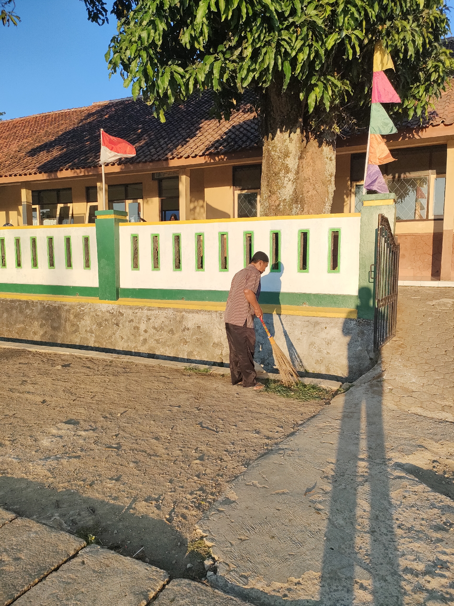 In Bojong, Indonesia, a person is sweeping the ground outside a building. The building has a tiled roof and a light-colored wall with green and yellow accents. A fence with rectangular openings runs along the front of the building. A large tree with lush green leaves stands near the building, and colorful flags are attached to a pole near the tree. The ground is a mix of dirt and concrete. The sun is shining, casting long shadows.