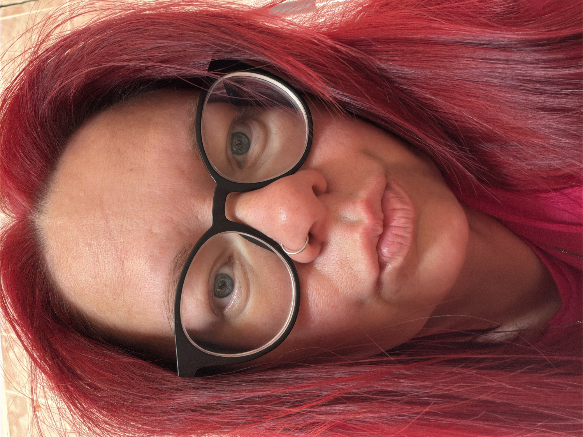 A close-up, slightly angled shot depicts a woman with vibrant red hair and fair skin. She is wearing black-rimmed glasses. A small silver ring is visible in her left nostril. The background is blurry and suggests a pink-colored garment. No elements visible in the frame allow to confirm any geographic location, including the city of Chaikovsky, Russia.