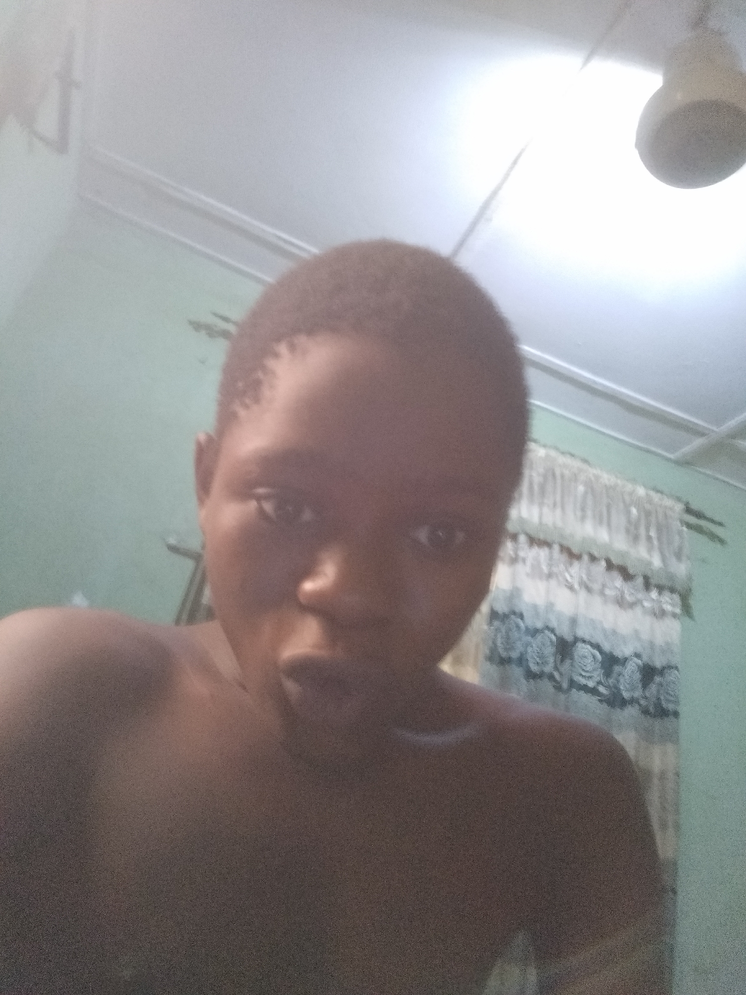 A close-up, slightly blurry image shows a person with dark skin and short hair, positioned indoors. The background reveals a light green wall, a curtain with a floral pattern, and a ceiling fixture. The location is Shangisha, Nigeria.
