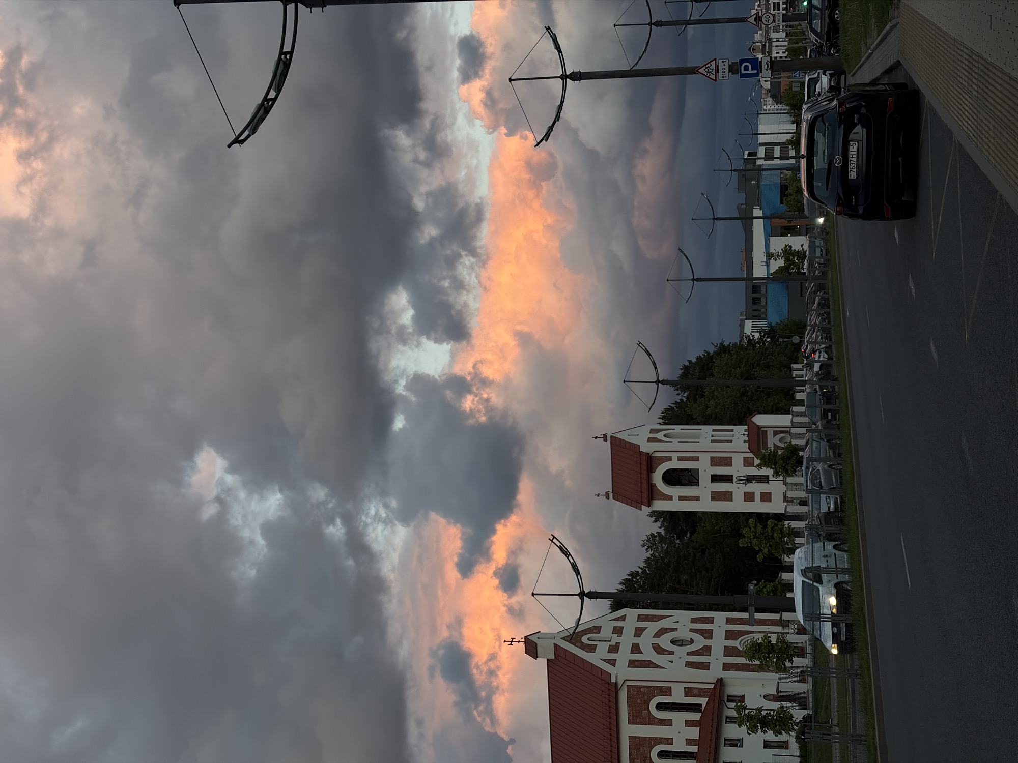 The image shows a street scene in Бараўлянскі сельскі Савет, Belarus. A road stretches along the right side of the frame, with a line of parked vehicles. On the left, there is a building with a distinctive white and red brick design, likely a church. Overhead, tram or trolleybus wires are suspended from poles. The sky is filled with dramatic, dark clouds with a patch of pink-tinged light.