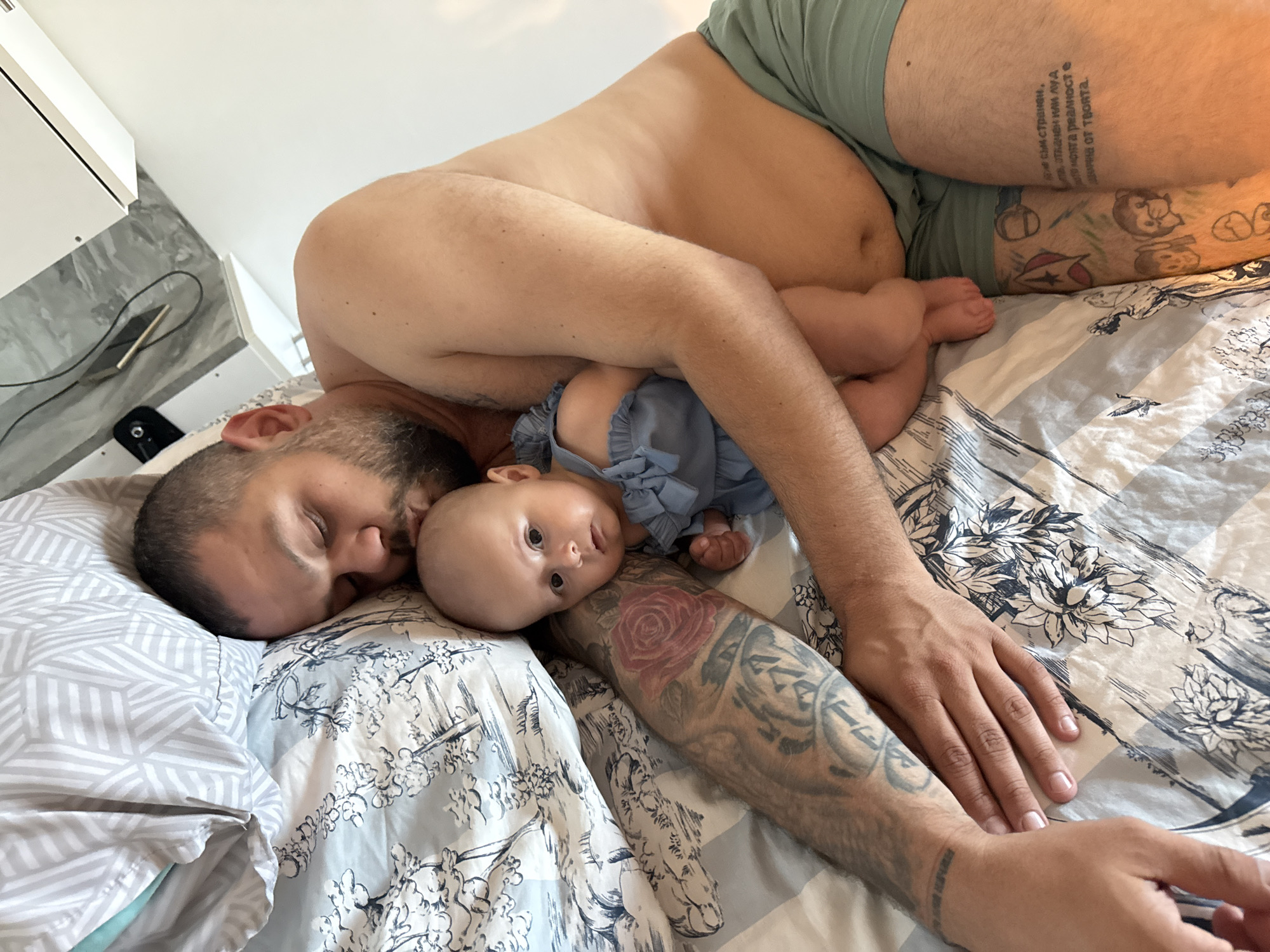 The image depicts a man and a baby lying on a bed. The man is on his side, with his eyes closed and his head resting on a pillow. He is holding the baby in his arms, and the baby is looking at the camera. The man has tattoos on his arm. The baby is wearing a blue outfit. The bed has patterned sheets. The background shows a white wall and a shelf. The location is identified as Plovdiv, Bulgaria.