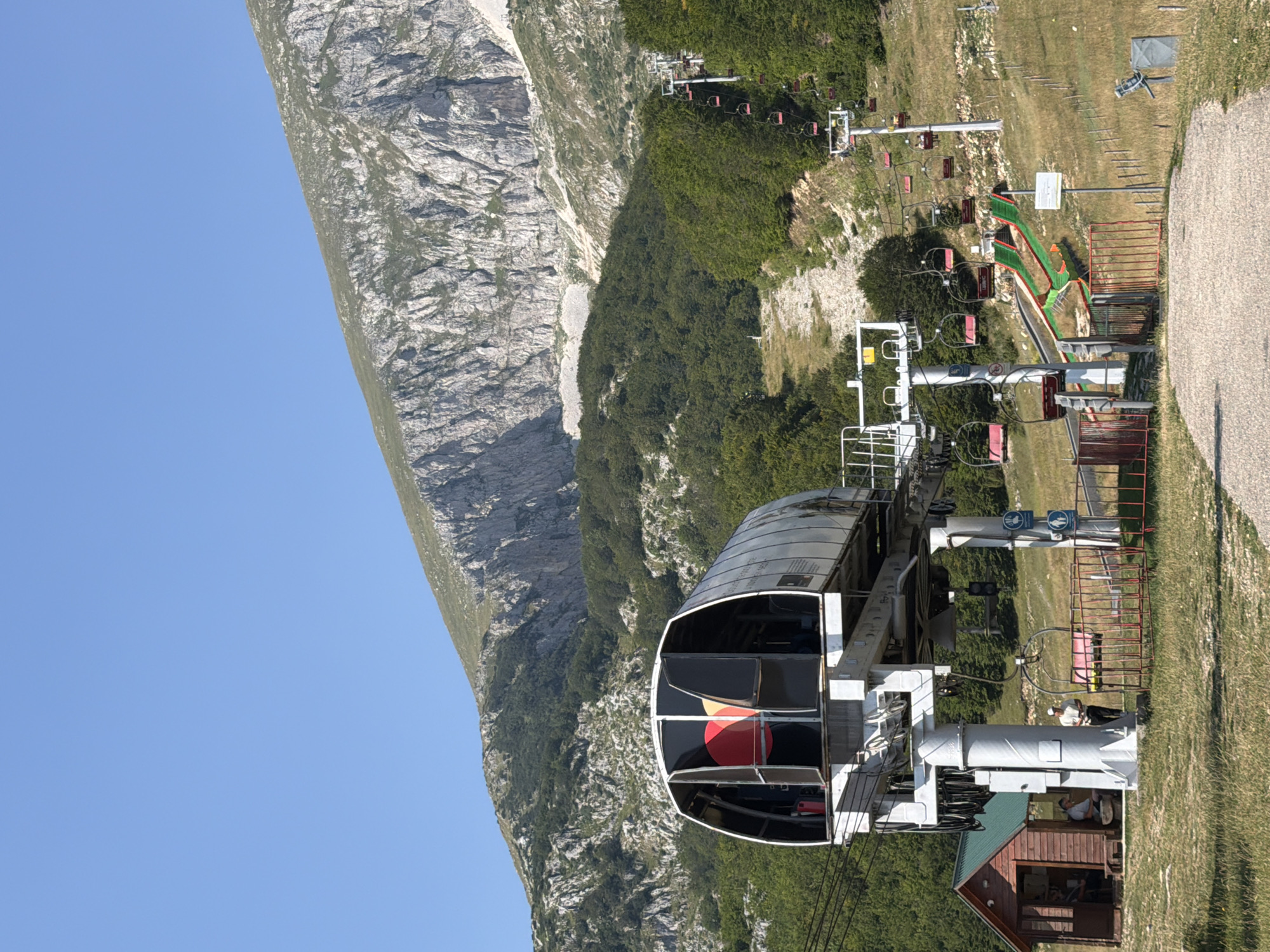 In Virak, Montenegro, a ski lift station sits at the base of a mountainside. The station features a large, enclosed cabin with red and orange circular motifs visible through the glass. The ski lift structure itself is supported by thick, white pillars, and numerous empty red chairlifts are suspended from cables ascending the grassy, tree-lined slope. Further up the mountain, the terrain becomes rocky and barren. A small wooden building with a green roof is situated to the left of the ski lift station, and two individuals are visible inside. Another person stands outside the building, near the ski lift. The sky is clear and blue.