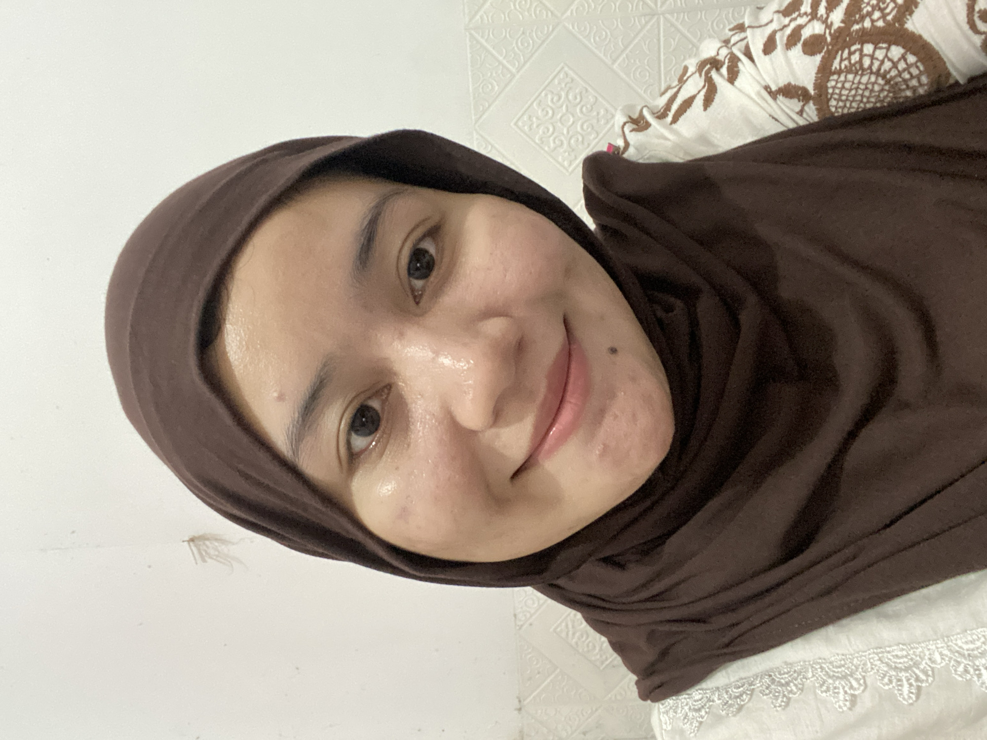 A close-up, head-and-shoulders selfie of a young Indonesian woman wearing a brown hijab is pictured. She has a light brown complexion and dark eyes, and she is smiling at the camera. Her skin has a slight sheen, indicating it may be oily or that she is wearing makeup. There are a few small blemishes visible on her cheeks and forehead. She is wearing a white top with brown embroidered detailing. The background consists of a white wall with a textured pattern and some faint markings, possibly from a previous insect or damage. The lighting is even and appears to be from a frontal source. There is no context provided to determine the specific location within Banjarbaru, Indonesia.