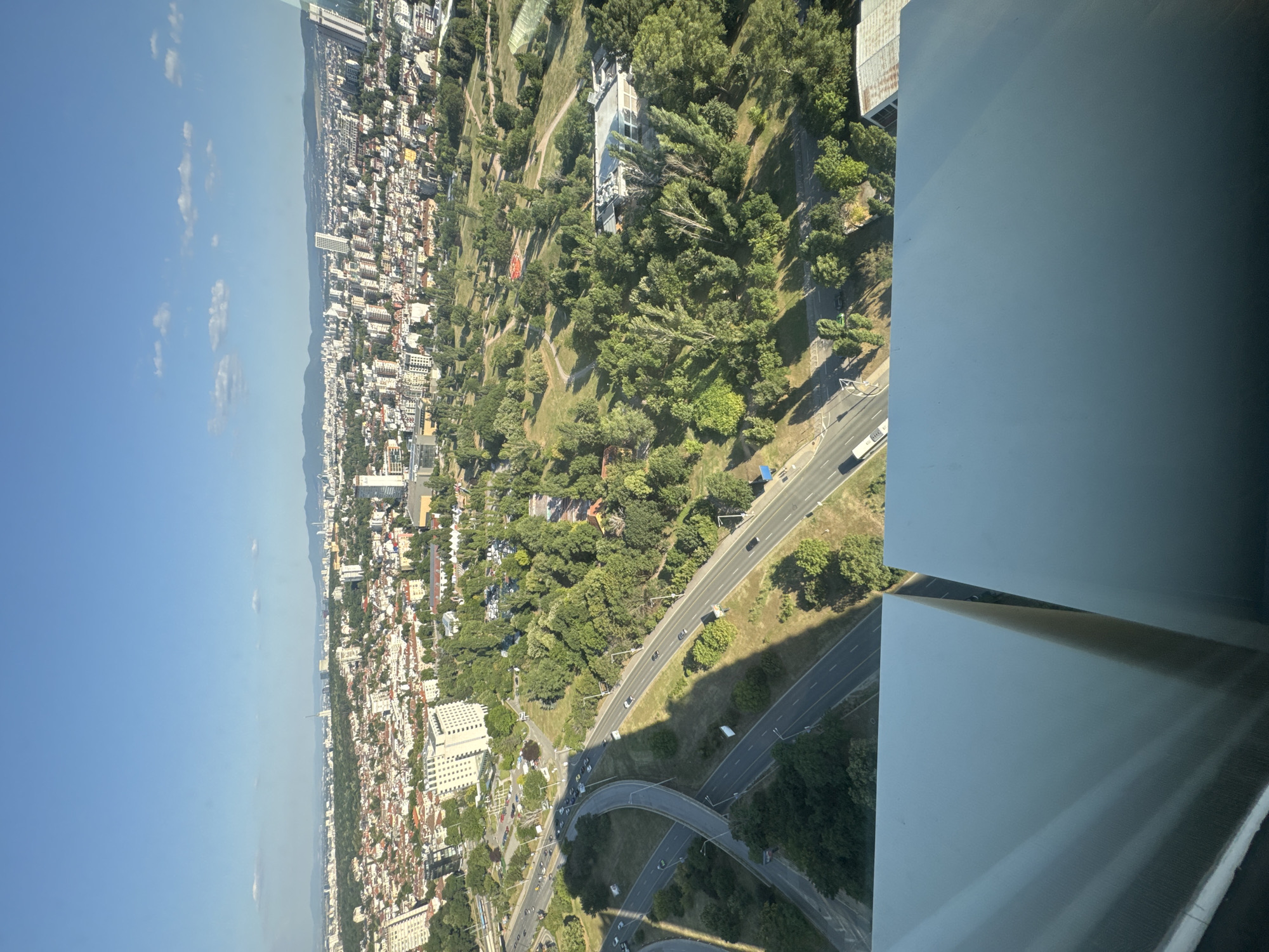 An aerial view captures a sprawling cityscape of Sofia, Bulgaria, under a clear blue sky dotted with a few white clouds. The foreground is dominated by the sleek, reflective surfaces of a modern building's exterior, appearing as large, angled planes that obscure the lower portion of the scene.
Beyond the building, the urban landscape unfolds with a mix of residential areas, commercial buildings, and extensive green spaces. Dense clusters of apartment buildings, many with terracotta-tiled roofs, transition into a large, verdant park. The park is characterized by numerous mature trees with lush green foliage, interspersed with pathways and open grassy areas. A prominent feature within the park is a modern, low-rise building with a light-colored facade and a rusted metal roof.
A complex network of elevated highways and roads snakes through the scene, with several cars and trucks visible on the asphalt. The infrastructure suggests a well-connected urban environment. In the distance, a more densely built-up area with taller skyscrapers rises against a backdrop of rolling hills and mountains that frame the city. The overall impression is one of a vibrant and well-established metropolis integrating natural elements within its urban fabric.
