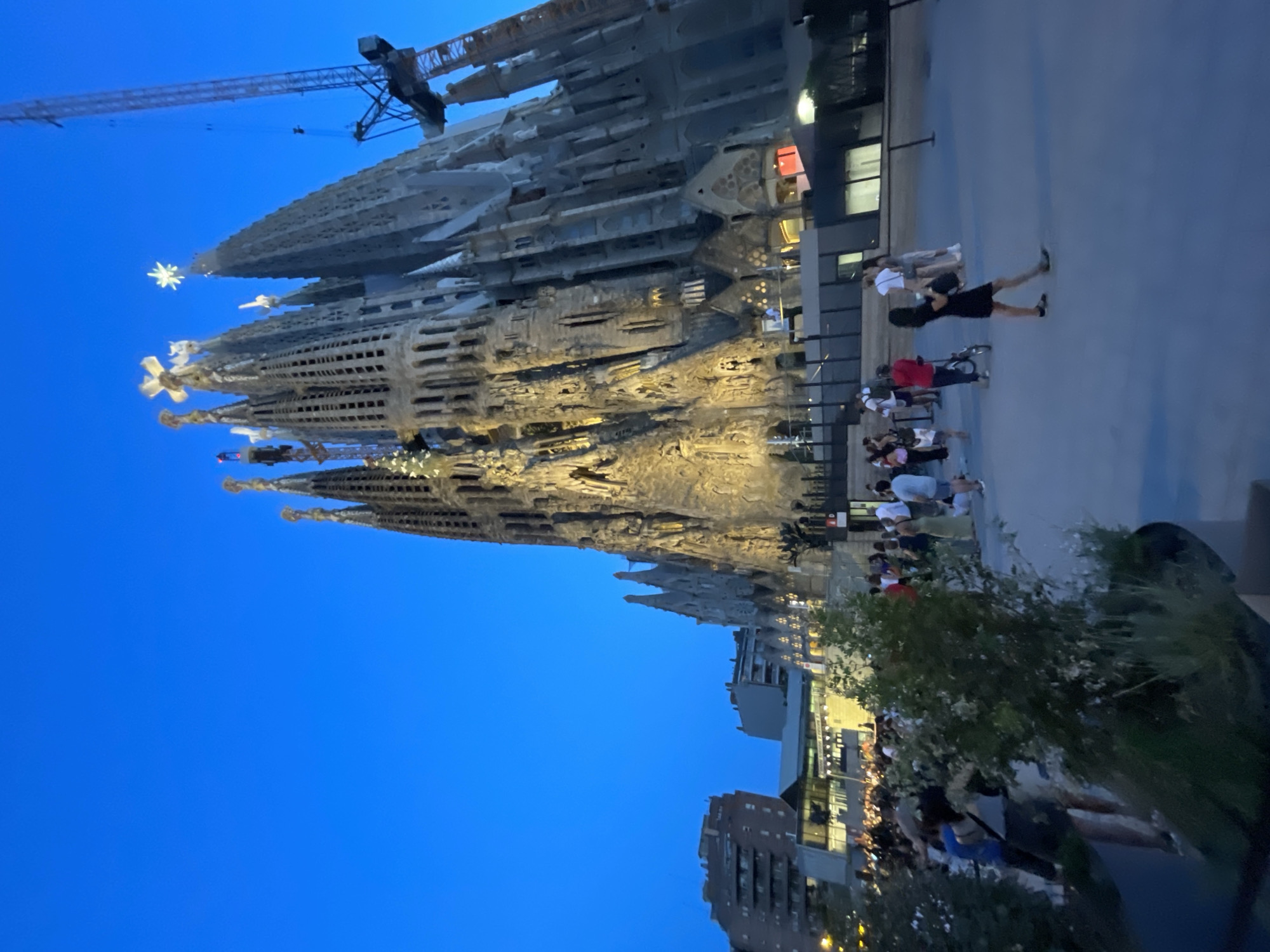 The image shows the Sagrada Familia basilica in Barcelona, Spain, at dusk. The basilica, a UNESCO World Heritage site, is illuminated and its spires are visible against a clear blue sky. A large crane is positioned next to one of the spires, indicating ongoing construction. In the foreground, a crowd of people are gathered in a plaza, some standing and talking, while others are walking or pushing a bicycle. The plaza is paved and features some greenery in planters. Buildings and infrastructure are visible in the background. The overall scene depicts a popular tourist destination in the evening.