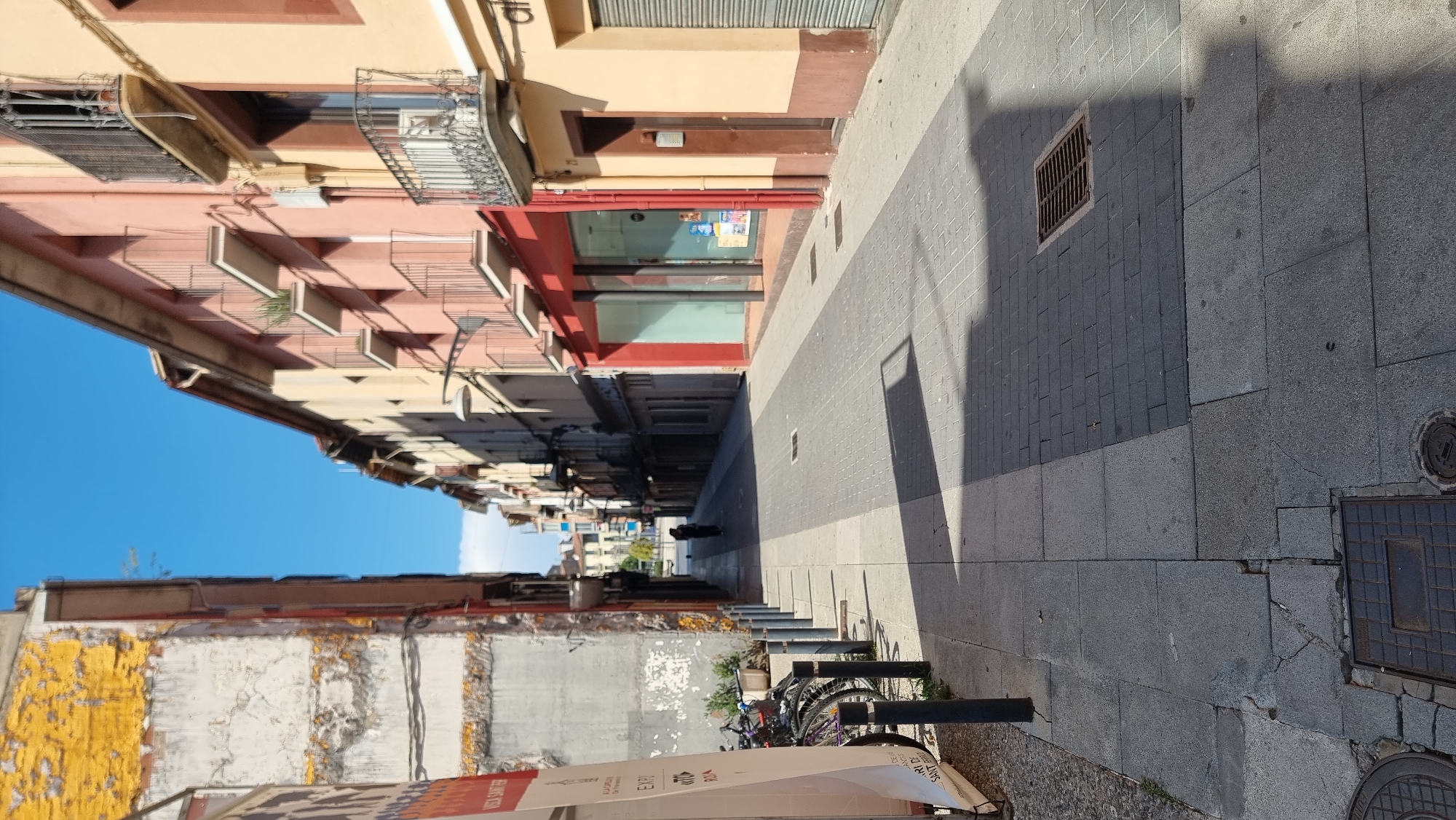 The image depicts a narrow street in Olot, Spain. The perspective is from the sidewalk, looking down the street. The buildings lining the street are of varying heights and architectural styles, with some featuring balconies. The street is paved with cobblestones and tiles, and there are shadows cast across the pavement, indicating a sunny day.
On the left side of the street, there is a building with weathered walls, partially covered by a large advertisement banner. Several bicycles are parked against the building. The right side of the street features shops with large windows and colorful facades, some with metal shutters. A lone figure is walking away from the camera in the distance, further down the street. The sky is clear blue. The overall scene portrays a typical European urban streetscape under bright daylight.