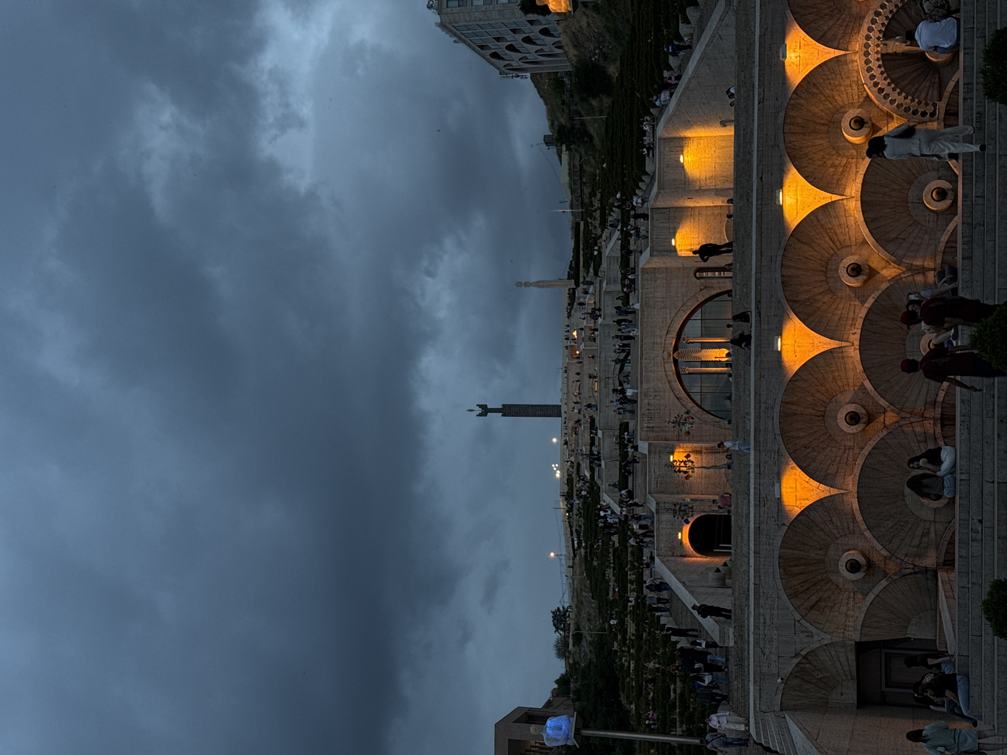 The image captures the Yerevan Cascade, a massive staircase with fountains and sculptures, at dusk or night. The perspective is from near the base of the cascade, looking upwards and across. The sky is overcast with heavy, dark gray clouds, suggesting an evening with low light or impending weather.
Numerous people are visible throughout the scene, scattered across the various levels, steps, and terraces of the cascade. Some individuals are walking up or down the stairs, while others are gathered in small groups, standing or sitting on the stone structures. The overall activity suggests a social evening, with people enjoying the public space.
The cascade itself is well-lit by artificial lights, casting warm, golden-orange glows on the stone surfaces, particularly highlighting the intricate, circular patterns of the fountains and decorative elements. This warm lighting creates a strong contrast with the dark, cool tones of the sky and unlit portions of the architecture. Buildings are visible on either side and behind the cascade, hinting at an urban setting.
In the distance, atop a hill, a tall statue, identifiable as the Mother Armenia statue, is silhouetted against the dark sky, serving as a prominent landmark. Another smaller, dark statue is also visible midway up the cascade. The design of the cascade includes numerous terraces with foliage, adding natural elements to the monumental stone structure. No visible text is discernible in the image. The scene is illuminated by the artificial lights, giving it a dramatic and inviting atmosphere despite the dark sky.