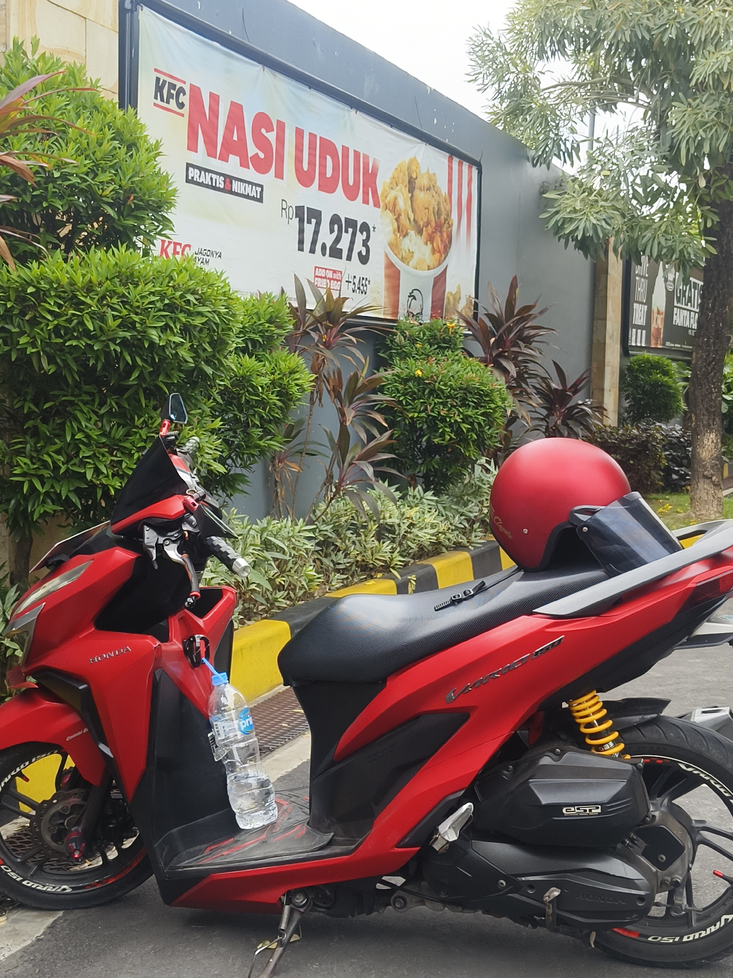 An outdoor scene in Denpasar, Indonesia, features a red Honda motorcycle parked in front of a KFC restaurant. The motorcycle is equipped with a red helmet and a water bottle. A large KFC billboard displaying "Nasi Uduk" (a traditional Indonesian rice dish) and the price of Rp 17,273 is visible in the background. Lush green bushes and trees surround the area, and a yellow curb marks the edge of the parking space. The ground is paved with asphalt.