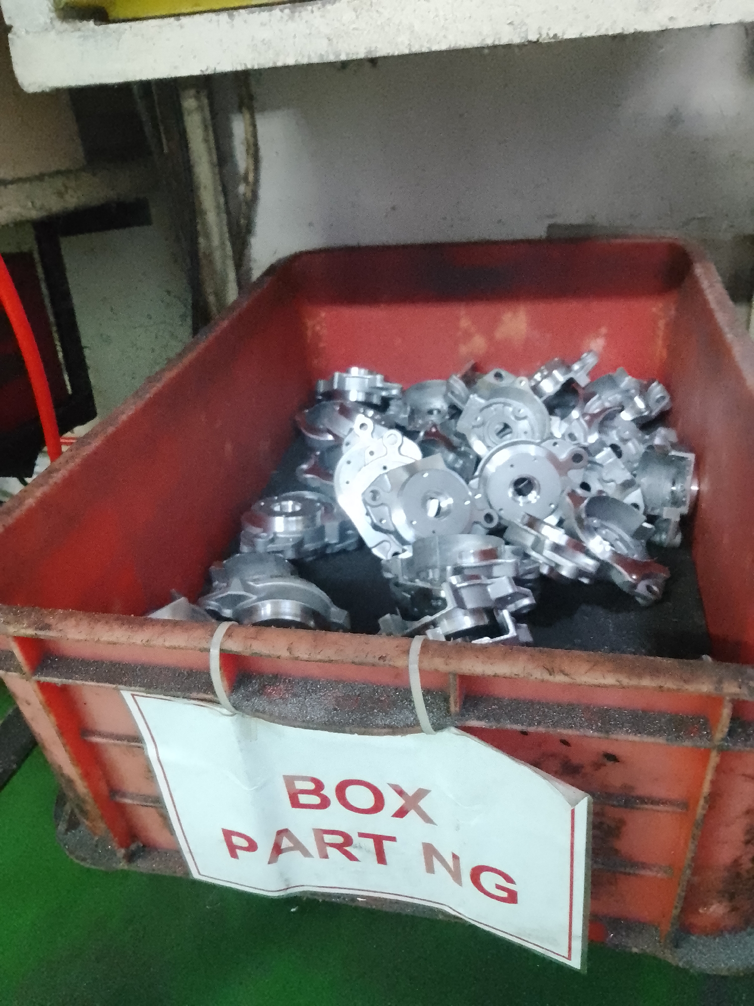 The image shows a red plastic bin filled with small, metallic parts. The bin is placed on a green surface, possibly a factory floor. The metallic parts appear to be machine components, possibly for an automotive or industrial application, given their intricate designs with holes and flanges. They are shiny and silver, suggesting they are made of aluminum or a similar metal.
Attached to the front of the bin is a white label with red lettering. The visible text on the label reads "BOX PART NG". This likely indicates that the contents are a box of parts, and "NG" could be an abbreviation for "No Good" or "Non-conforming", suggesting these parts are either scrap, awaiting inspection, or have failed a quality check.
The setting appears to be an industrial or manufacturing environment, as suggested by the bin, the parts, and the overall utilitarian appearance of the surroundings. The lighting is artificial, casting some shadows and highlighting the metallic sheen of the parts. There are no people visible in the image. The image does not provide any specific time of day or weather cues, but the overall impression is one of an active industrial workspace. The location context of Ciantra, Indonesia, further supports the industrial setting.