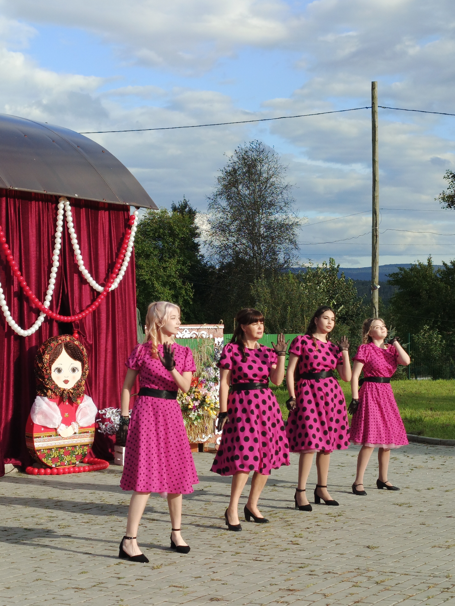 Four young women wearing matching bright pink dresses with black polka dots stand in a line on a paved outdoor surface, likely in Medvedyovka, Russia. They are dressed in vintage style with black belts, black gloves, and black low-heeled shoes. Their hair is styled, and they are all making a gesture with their hands, suggesting they are performing or posing.
To their left, there is a large, decorated cardboard cutout of a Matryoshka doll, a traditional Russian nesting doll. Behind the women and the doll is a stage or backdrop area draped with deep red fabric, adorned with strings of white and red balloons. Further behind the stage, there are trees, a grassy area, and rolling hills in the distance under a partially cloudy sky. A wooden utility pole with electrical wires runs vertically in the right side of the frame, indicating an outdoor, possibly rural or semi-rural, setting. The lighting suggests it is daytime, possibly late afternoon, given the warm tones.