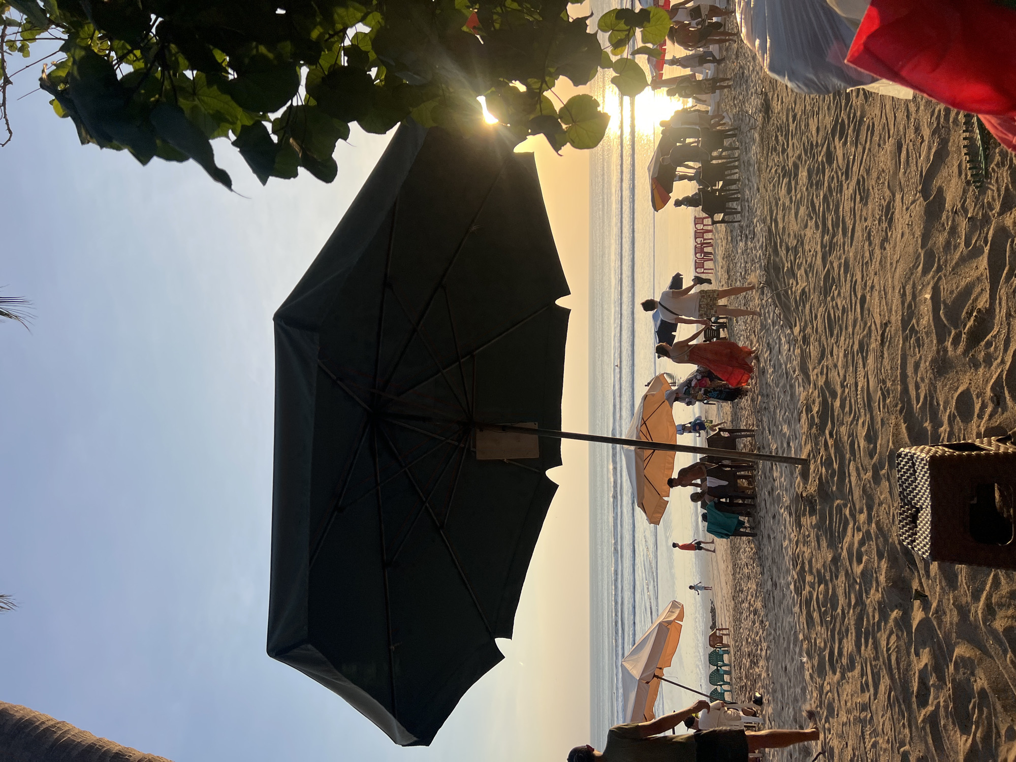 A beach scene in Kuta, Indonesia, at sunset. The foreground features a large, dark umbrella casting a shadow over a sandy beach. To the left, a person stands with their back to the camera. In the middle ground, several smaller umbrellas are set up, with people gathered around tables and chairs. The ocean is visible in the background, with gentle waves and the sun setting over the horizon, casting a warm glow. A tree with large leaves is visible in the upper right corner, partially obscuring the sun. The overall atmosphere is relaxed and tropical.
