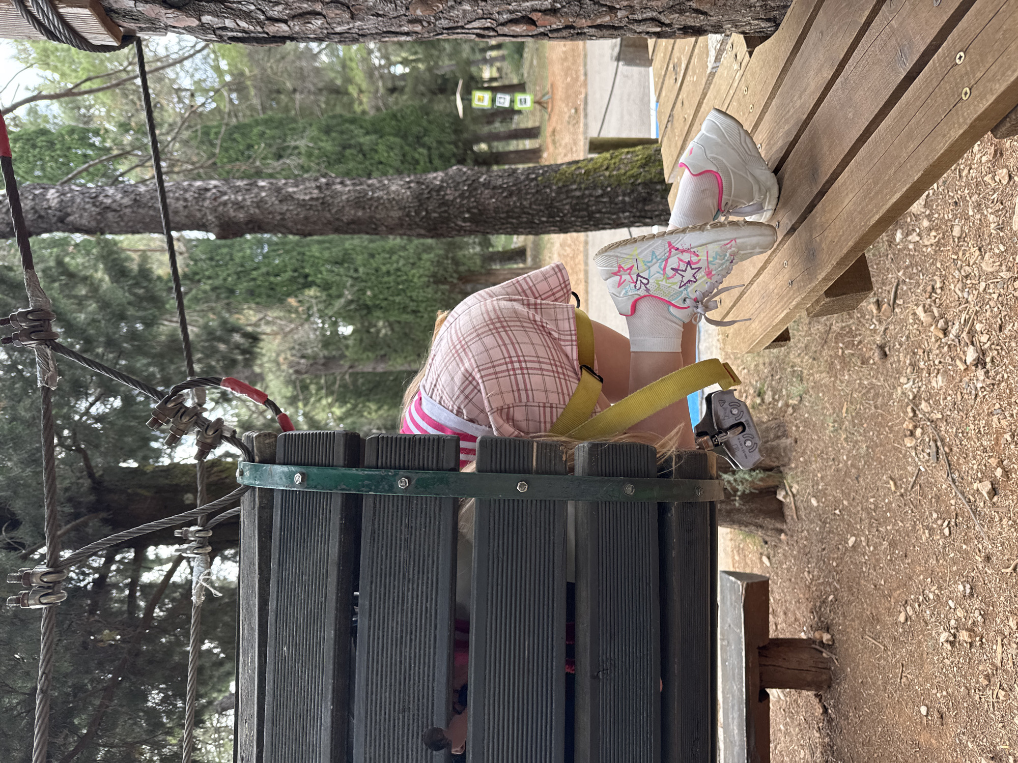 A young girl is suspended in a safety harness at an outdoor adventure park in Podgorica, Montenegro. She is wearing a plaid sun hat, a pink and white striped shirt, white socks, and white sneakers decorated with stars. The girl is positioned between a gray, vertical wooden structure and a wooden platform, secured by cables and hardware. The surrounding environment includes trees, and brown dirt ground.