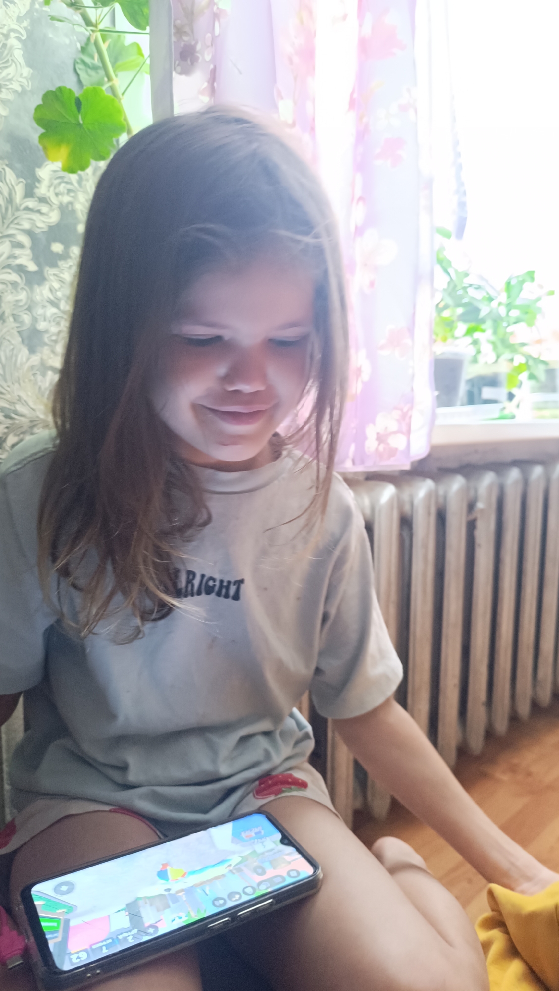 A young girl with long brown hair, wearing a light gray t-shirt with the word "RIGHT" printed on it and patterned shorts, sits on the floor, looking down at a smartphone she is holding in her lap. The phone displays a colorful game with cartoonish graphics. The background shows a radiator against a wall, and a window with sheer, floral curtains and greenery visible outside. The lighting suggests it is daytime, possibly near a window. This scene is located in the city of Karsha, Russia.