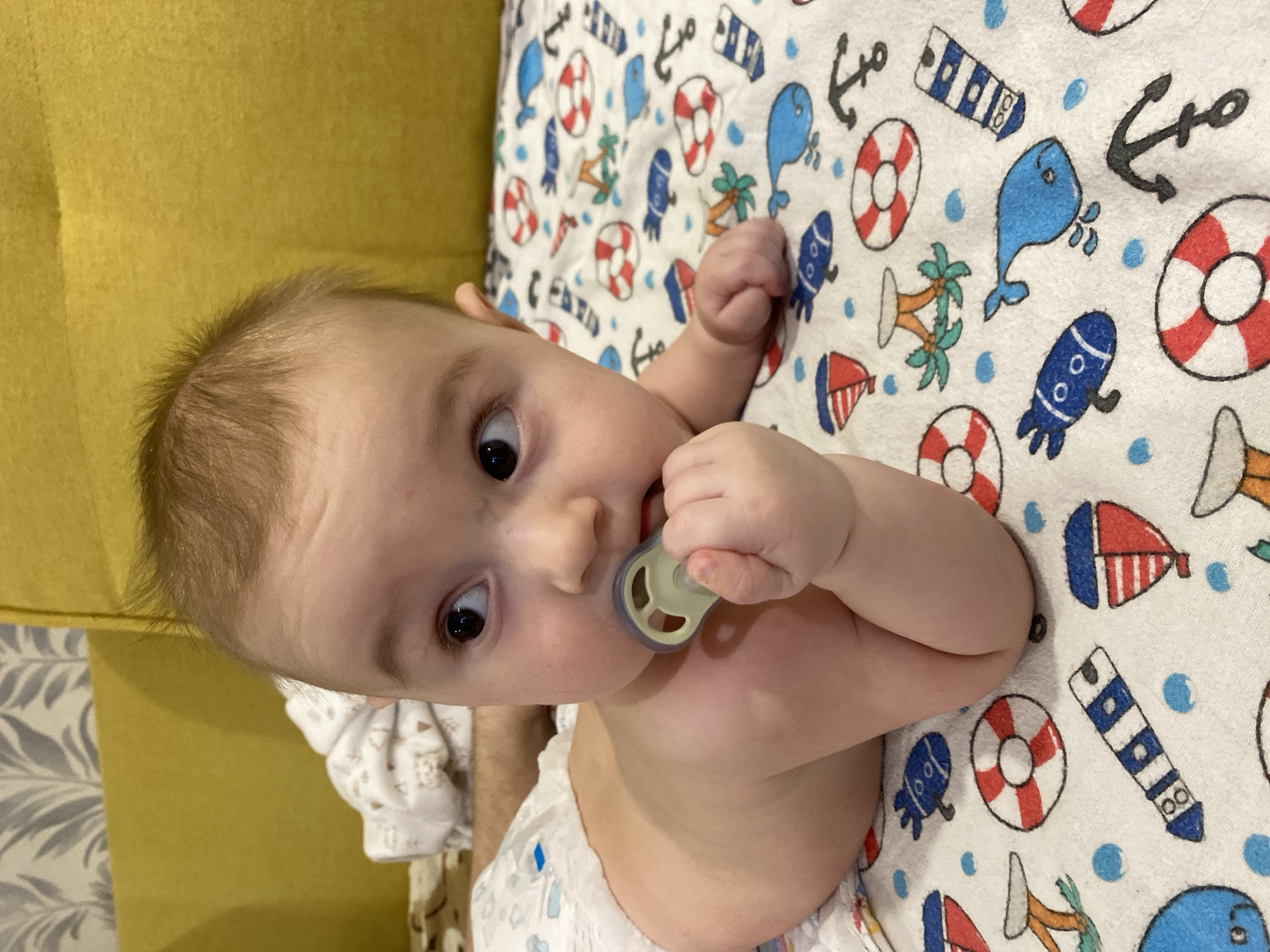 A baby with a pacifier is lying on a patterned blanket. The blanket features nautical designs including anchors, life preservers, whales, palm trees, sailboats, and blue dots. The baby's right hand is raised near the pacifier. A yellow sofa is visible in the background. The location is Chaikovsky, Russia.