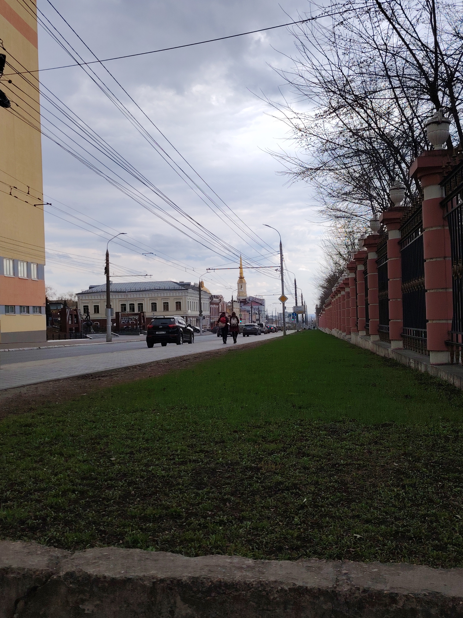 The image captures an urban street scene in Izhevsk, Russia, on an overcast day, likely during early spring given the bare tree branches and sparse, newly grown green grass.
In the foreground, a strip of green grass with emerging growth occupies the lower right, bordered by a dark concrete curb. Along the right side of the image, a long, ornate fence features regularly spaced pillars of a reddish-brown color, topped with decorative white urns, and black metal railings between them. Behind this fence, numerous bare tree branches are visible against the cloudy sky.
The main subject is a street running diagonally from the lower left towards the upper right. On the left side of the street, a tall, light-colored building with multiple windows stands prominently. Further down the street, a row of older-style buildings with light facades and dark roofs can be seen. In the distance, a distinctive building with a golden spire, possibly a church or cathedral, is visible. Numerous overhead power and trolleybus wires crisscross the sky, connecting to poles along the street.
On the paved street, a black SUV, likely a Hyundai Creta, drives away from the viewer in the left lane. On the sidewalk to the right of the street, two pedestrians, appearing to be women, walk in the same direction. Other vehicles are visible further down the road, indicating active traffic. Streetlights are also part of the urban infrastructure.
The weather is cloudy with a gray, overcast sky. No explicit text is clearly legible on buildings or signs. The scene depicts a typical bustling city environment on a cool, bright but sunless day.