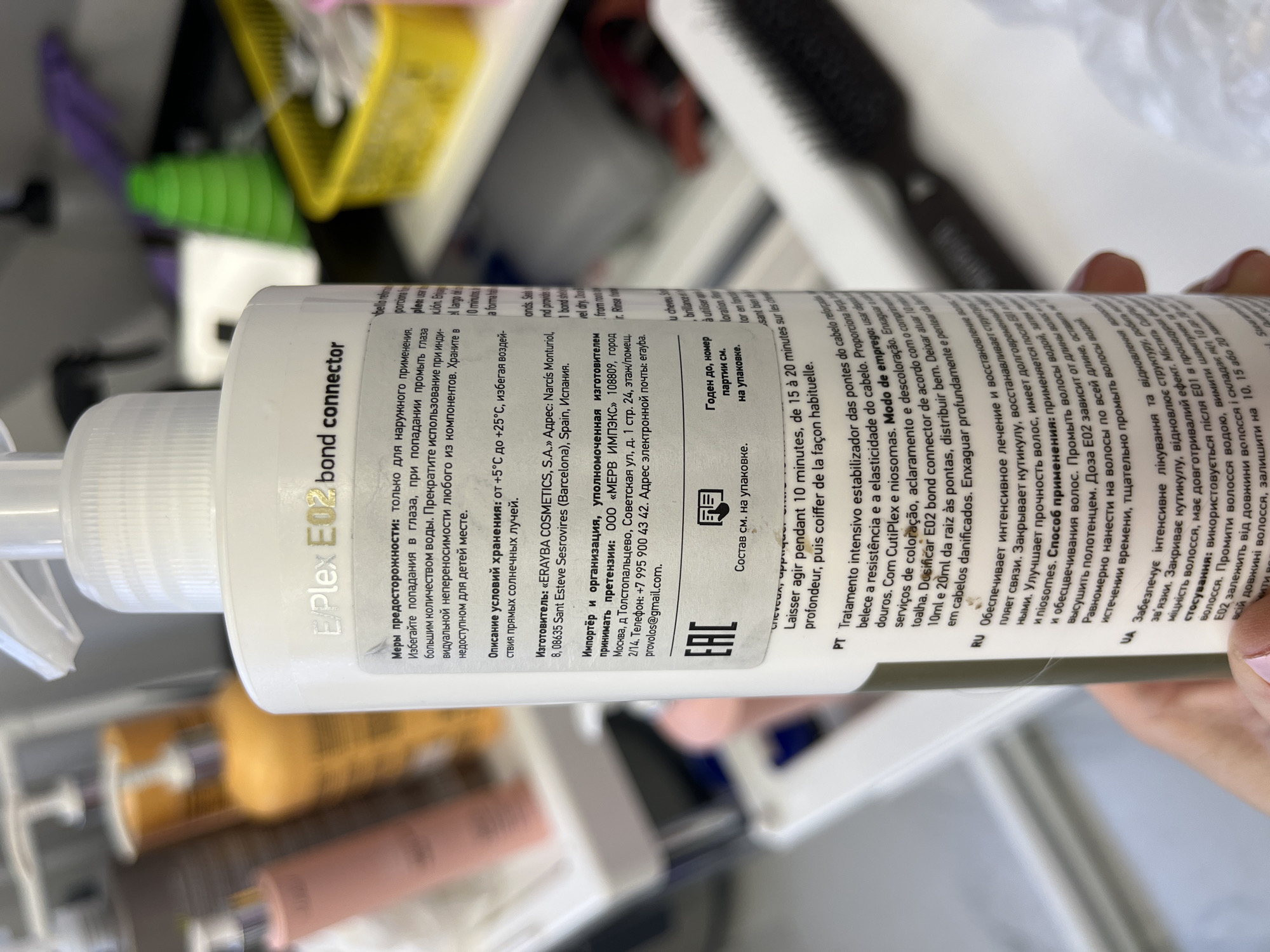 A person's hand is holding a white spray bottle of hair care product, specifically "EPlex E02 bond connector." The bottle's label is prominent, featuring detailed information in multiple languages, primarily Russian, Spanish, Portuguese, and Ukrainian.
Visible text on the label includes:
- Product name: "EPlex E02 bond connector"
- Warnings: "Меры предосторожности: только для наружного применения. Избегайте попадания в глаза..." (Precautions: for external use only. Avoid contact with eyes...)
- Storage conditions: "Описание условий хранения: от +5°С до +25°С, избегая воздействия прямых солнечных лучей." (Storage conditions: from +5°C to +25°C, avoiding direct sunlight.)
- Manufacturer: "Изготовитель: «ERAYBA COSMETICS, S.A.»" located in Sant Esteve Sesrovires (Barcelona), Spain.
- Importer: "Импортёр и организация, уполномоченная изготовителем принимать претензии: ООО «МЕРВ ИМПЭКС»" in Moscow, Russia, with an address, phone number (+7 995 900 43 42), and email (erayba.provolos@gmail.com).
- Instructions for use: Descriptions in Russian, Portuguese, and Ukrainian mention intensive treatment, strengthening hair, restoring structure, improving elasticity, and ingredients like CutiPlex and niosomes. Application instructions suggest applying after E01, drying with a towel, distributing evenly, leaving for 10-20 minutes, then rinsing.
- Batch and expiry information: "Годен до, номер партии см. на упаковке." (Best before, batch number see on packaging.)
- "Состав см. на упаковке" (Ingredients see on packaging) and "EAC" marking.
The background is out of focus, showing what appears to be shelves or containers filled with various beauty or personal care products, including yellow and brown bottles, and some green and purple organizational bins, suggesting an indoor setting like a salon, bathroom, or storage area. The lighting is bright.