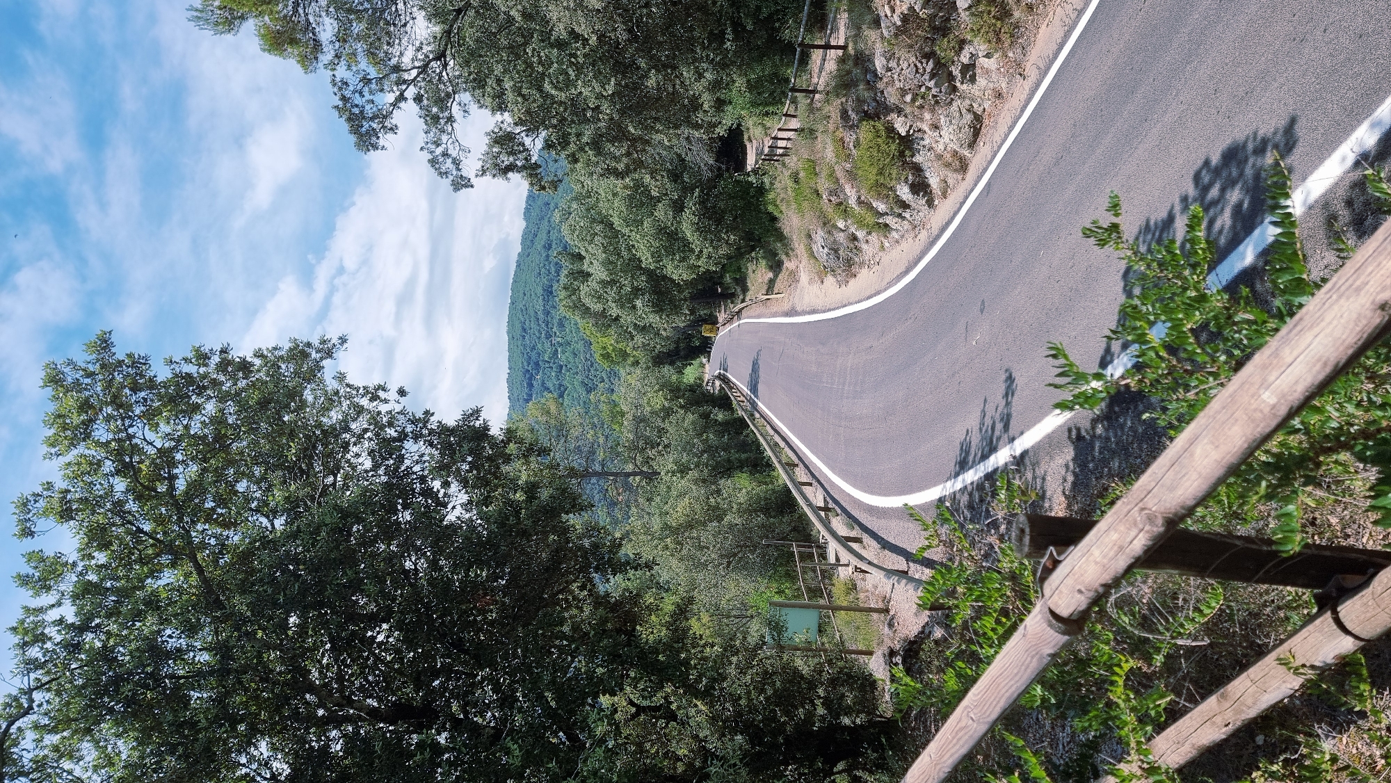 The image depicts a winding asphalt road with white lane markings, curving through a forested hillside. The road is bordered by a wooden guardrail and lush green vegetation. In the distance, rolling hills covered in dense trees are visible under a partly cloudy blue sky. The scene suggests a rural or natural setting. The location is identified as Tortellà, Spain. There are no people or animals visible in the image. The overall atmosphere is serene and natural.