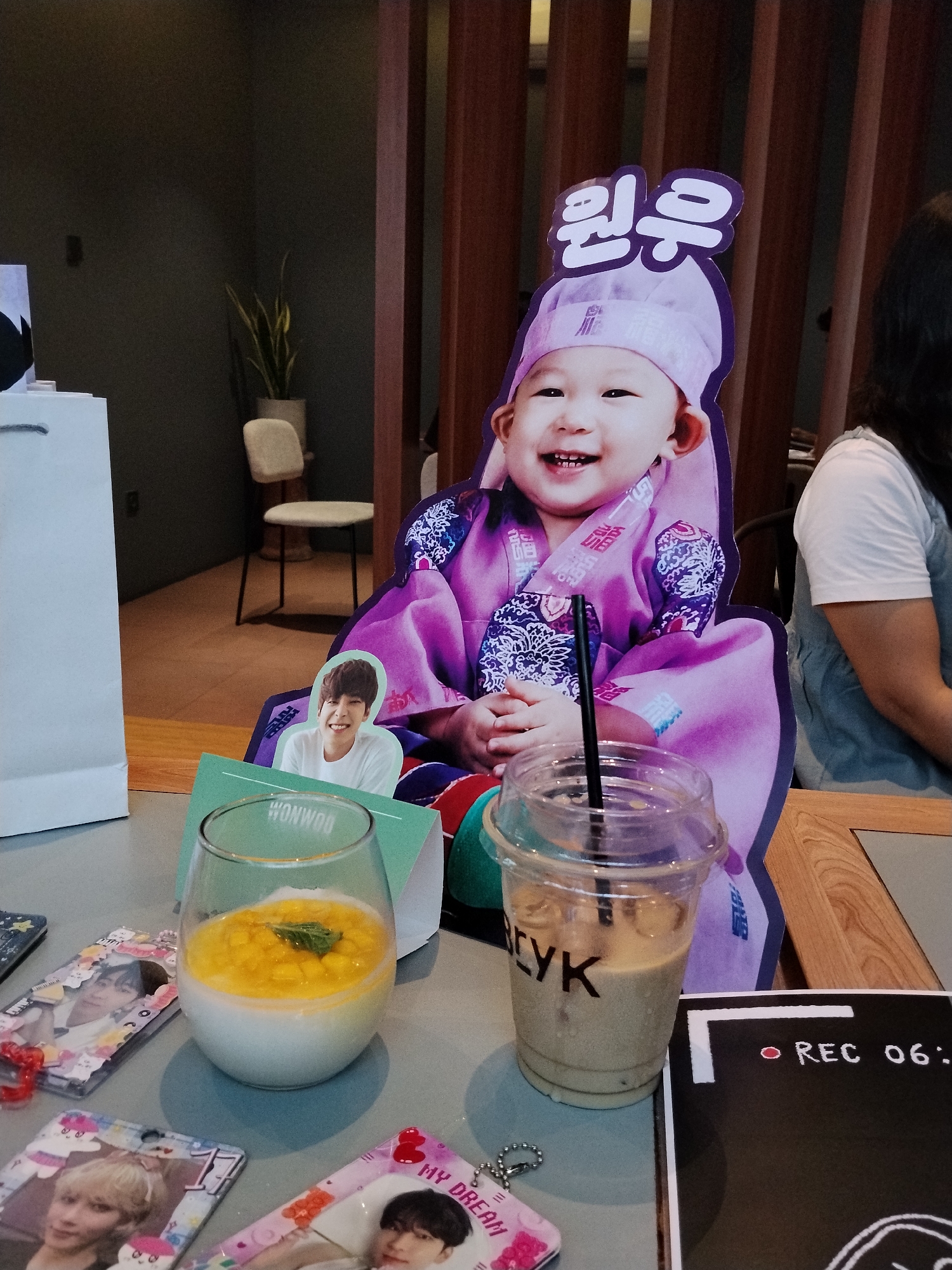 In the city of Serang, Indonesia, this image captures a close-up view of a table at what appears to be a cafe. The foreground is dominated by a cardboard cutout of a smiling baby in a traditional Korean hanbok, with Korean text at the top. To the left of the baby cutout, there is a smaller cutout of a young man. In front of these cutouts are several key objects. A glass contains a white dessert topped with diced yellow fruit and a sprig of mint. Next to it is a clear plastic cup filled with a light brown beverage and a straw. Scattered around these drinks are several small, rectangular items featuring images of young men, resembling photocards or keychains. A black board with a red circular "REC" icon and the text "06:" is visible to the right, suggesting a recording device or theme. In the background, a woman with dark hair is partially visible, wearing a white t-shirt and a grey apron-like garment. The environment appears to be indoors, with wooden furniture and wall paneling visible.