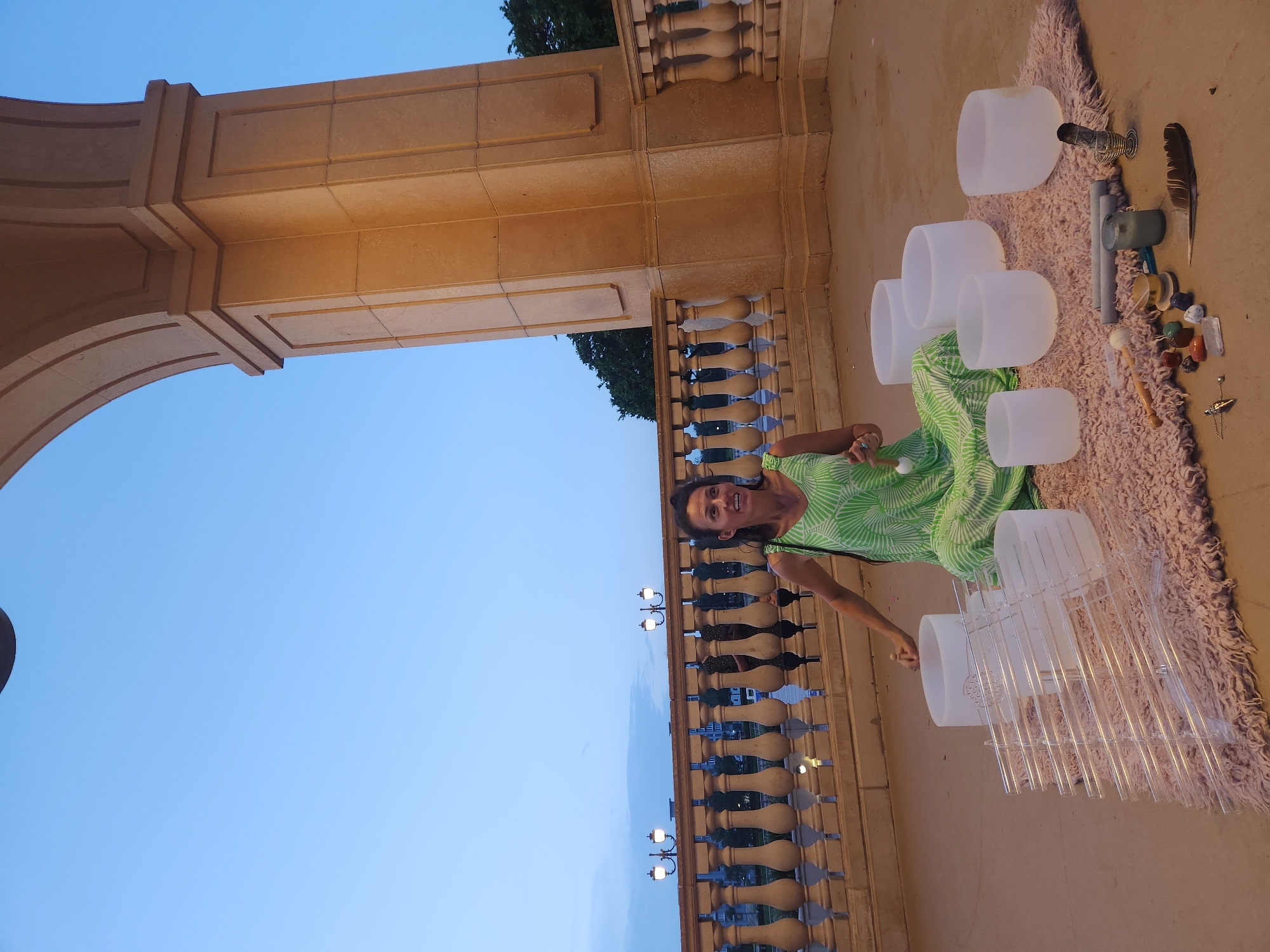 A woman in a green and white patterned dress sits cross-legged on a pink rug in front of a row of crystal singing bowls arranged on the ground. She holds a mallet in her right hand, poised over one of the bowls. To the left of the bowls are a series of clear glass tubes arranged on a rack. The scene appears to be an outdoor setting, possibly a patio or balcony, with an ornate stone archway and balustrade visible in the background. The sky is a pale blue, suggesting it is either early morning or late evening. The location is identified as Altamonte Springs, United States of America.