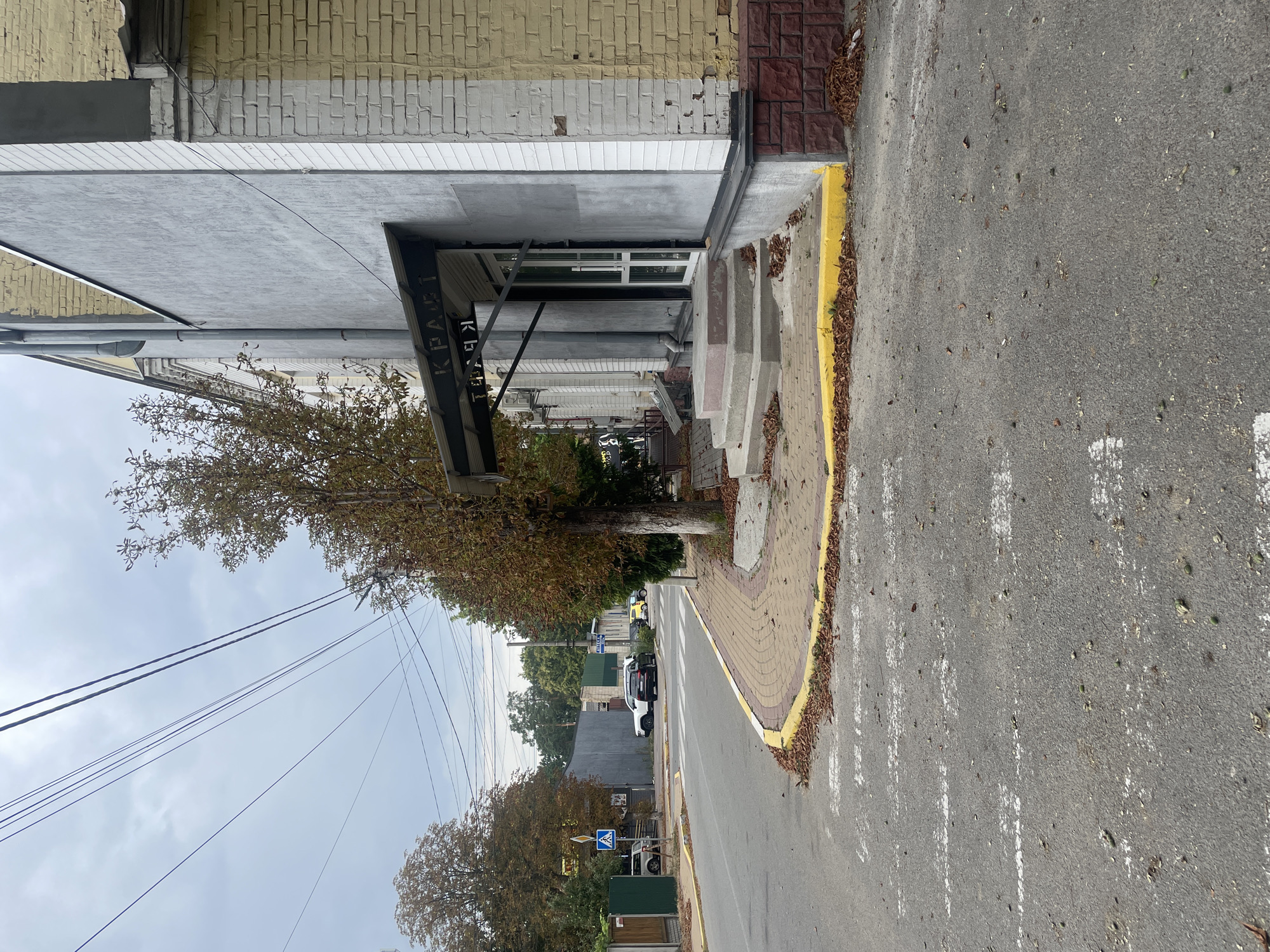 The image shows a street scene in Vorzel, Ukraine, with a focus on a building entrance. The building has a grey facade with a yellow brick upper section. Above the entrance, a black awning with the Cyrillic word "КРАФТ" (Kraft) is visible. The entrance itself consists of glass doors. The building is situated next to a sidewalk with yellow and brown paving stones. A street with a crosswalk marked by white stripes runs in front of the building. Several cars are parked on the street, including a white SUV and a black sedan. Trees with autumn foliage are present on either side of the scene. The sky is overcast. The overall impression is that of a quiet, residential or commercial street in a suburban setting during the autumn season.