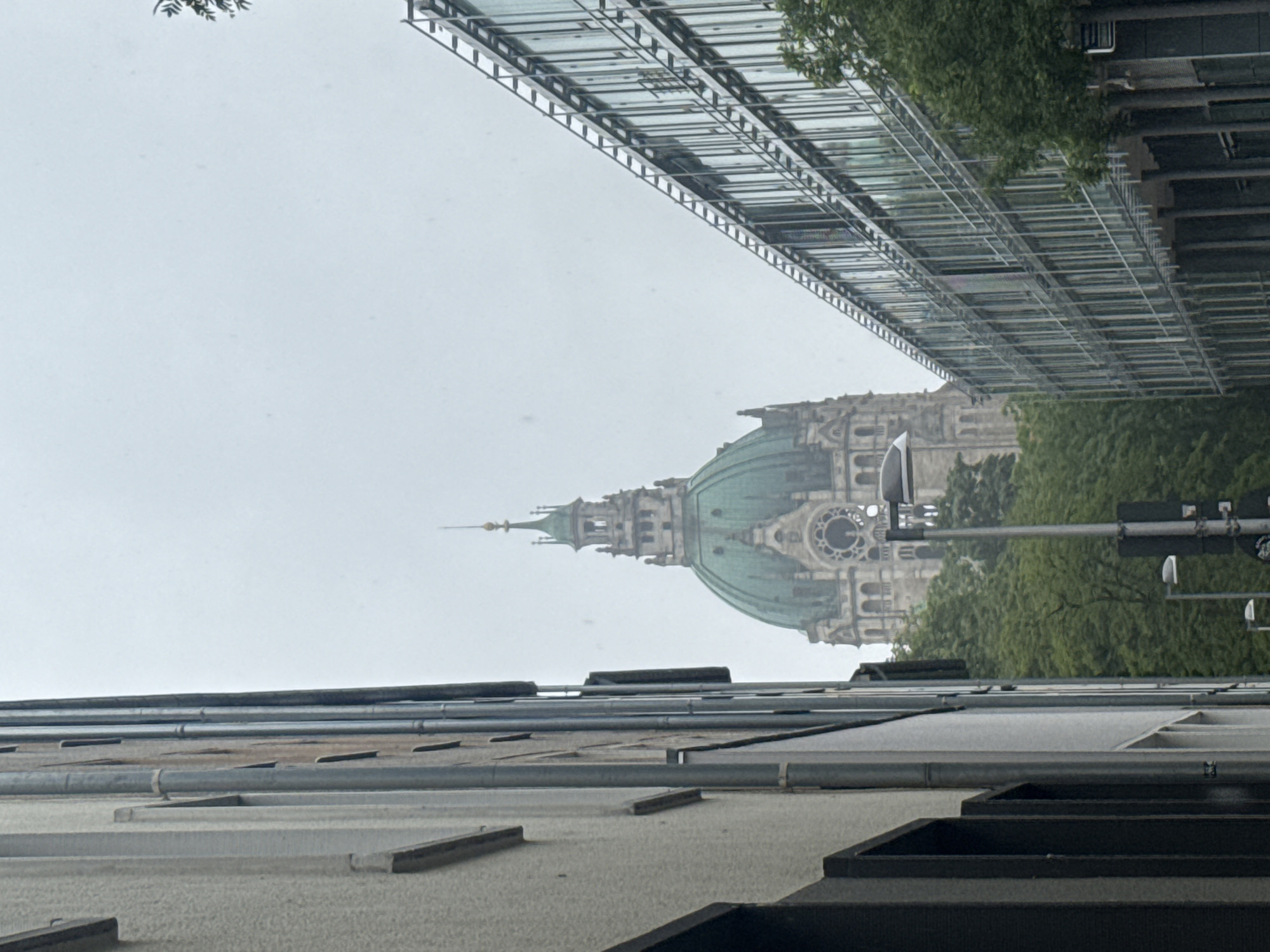 The image shows a street view in a city. On the left, there is a building with a light-colored textured facade and dark vertical window frames. On the right, there is a modern building with a glass facade and horizontal metal elements. In the background, a large dome-shaped building with a green roof and intricate architectural details is visible, surrounded by lush green trees. The sky is overcast and a street lamp and traffic signals are present in the foreground.