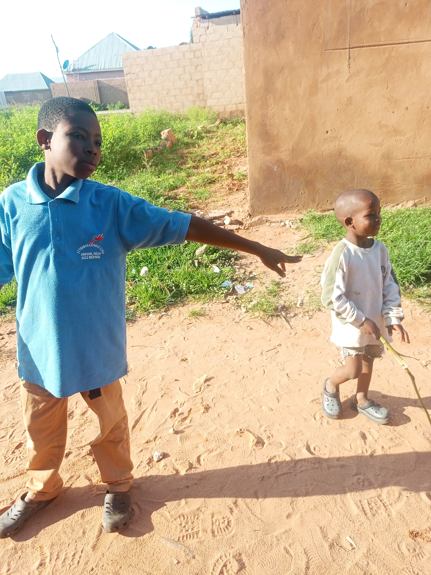 The image depicts two young boys in an outdoor, urban environment. The boy on the left, wearing a blue polo shirt with an emblem and tan pants, is positioned in the foreground, facing right and gesturing with his left arm. He is wearing dark clogs. The boy on the right, in the middle ground, wears a light-colored long-sleeved shirt and shorts, and also has dark clogs. He is holding a thin green stick. The ground is sandy and appears to be a dirt path. In the background, there are buildings constructed of brick and stucco, along with green vegetation. The scene suggests a sunny day with distinct shadows cast by the boys and objects. Based on the provided context, the location is Tempure, Nigeria.