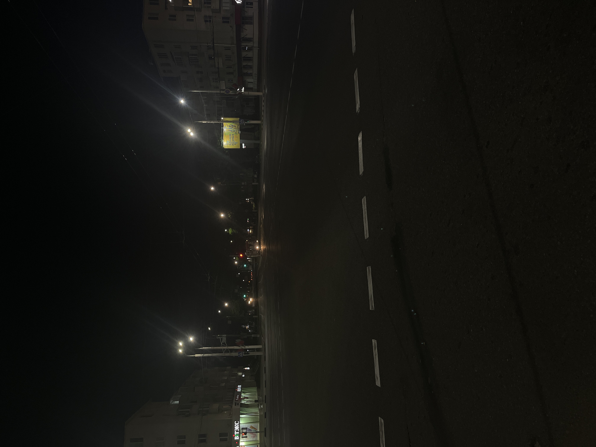 The image depicts a nighttime view of an intersection in Vitsebsk, Belarus. Streetlights illuminate the asphalt road and sidewalks, casting long shadows. On the left side of the image, a multi-story apartment building with illuminated windows stands adjacent to a store with a brightly lit sign. Across the intersection, another apartment building is visible. Traffic lights are illuminated, indicating a red light for traffic in one direction and a green light for traffic in another. A vehicle is seen in the distance, its headlights on. A bus stop sign is also present on the right side of the road. The scene is quiet and deserted, with no people visible in the frame.