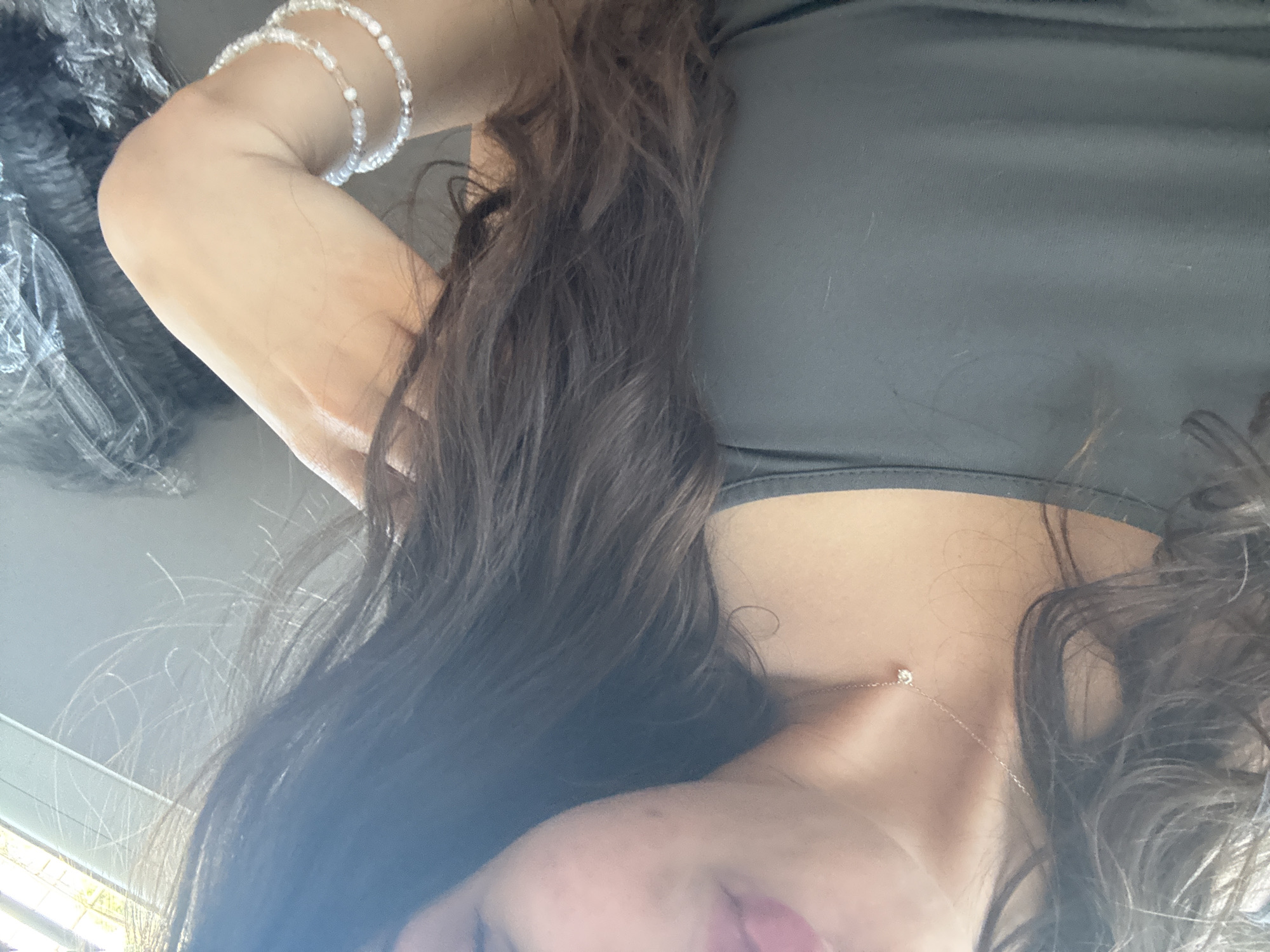 The image is a close-up, waist-up shot of a person, likely a woman, indoors, with a portion of her face visible. She has dark, wavy hair that falls across her shoulder and chest. She is wearing a dark, sleeveless tank top. Around her neck, she wears a delicate silver necklace with a small, clear pendant. On her right arm, she has two bracelets, one thinner and one wider, both made of small, iridescent beads. The background is out of focus and appears to be an indoor setting with a window or bright light source visible on the right side. There is also a dark, crinkled object partially visible on the far right. The image does not provide any information or context related to the city of Dibich, Bulgaria.