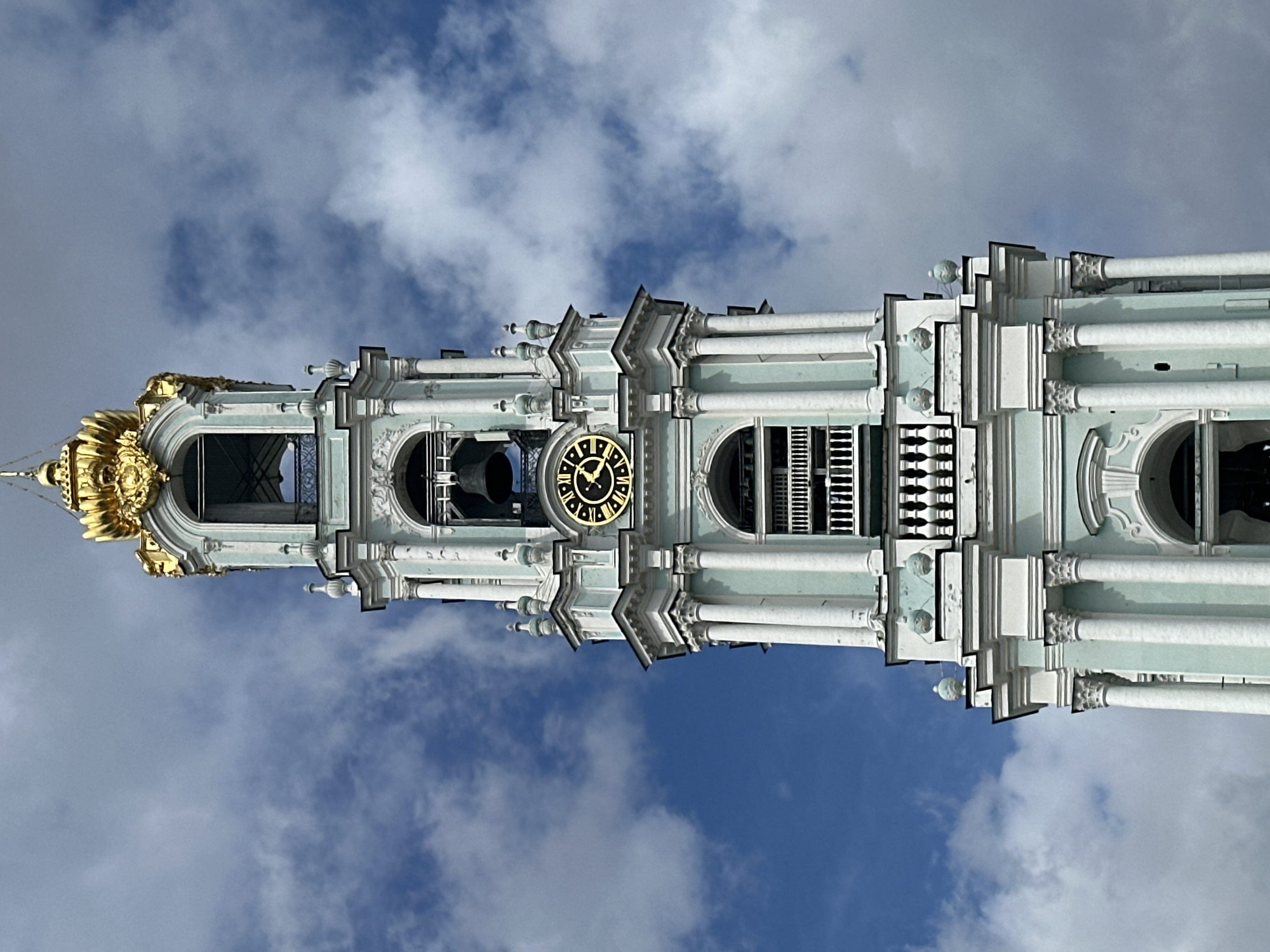 A low-angle, full shot captures the upper portion of a pale teal and white bell tower, topped with a bright gold, ornate crown-like structure. The tower features multiple levels with arched windows, decorative columns, and balconies. A prominent clock face with Roman numerals and black hands is visible on one of the mid-levels. A large, dark bell hangs in an opening above the clock. The tower is set against a bright blue sky filled with scattered white clouds.