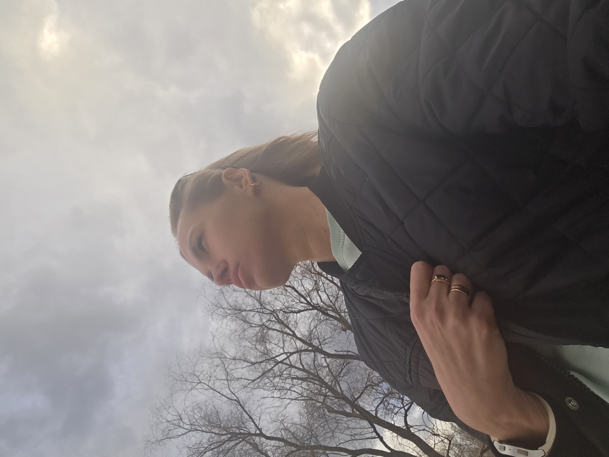 This low-angle, profile shot captures a person outdoors against a backdrop of a cloudy sky and bare trees. The subject, appearing to be female, is positioned in the upper left portion of the frame, facing right, with her left side visible. She wears a black quilted jacket over a light blue or mint green garment. Her light brown hair is pulled back, and she has a small gold hoop earring. Her expression is somewhat pensive or introspective, with her gaze directed slightly downwards and her lips gently pursed.
Her right hand, adorned with two gold rings (one plain band, one with a dark stone), rests on her jacket near her chest. A white smartwatch is visible on her left wrist.
The background features a vast, overcast sky with a bright, diffused light source, indicative of the sun behind clouds. In the lower right, the stark, leafless branches of deciduous trees suggest the season is late autumn, winter, or early spring. This natural setting is located in Noviy, Russia. No visible text is present in the image.