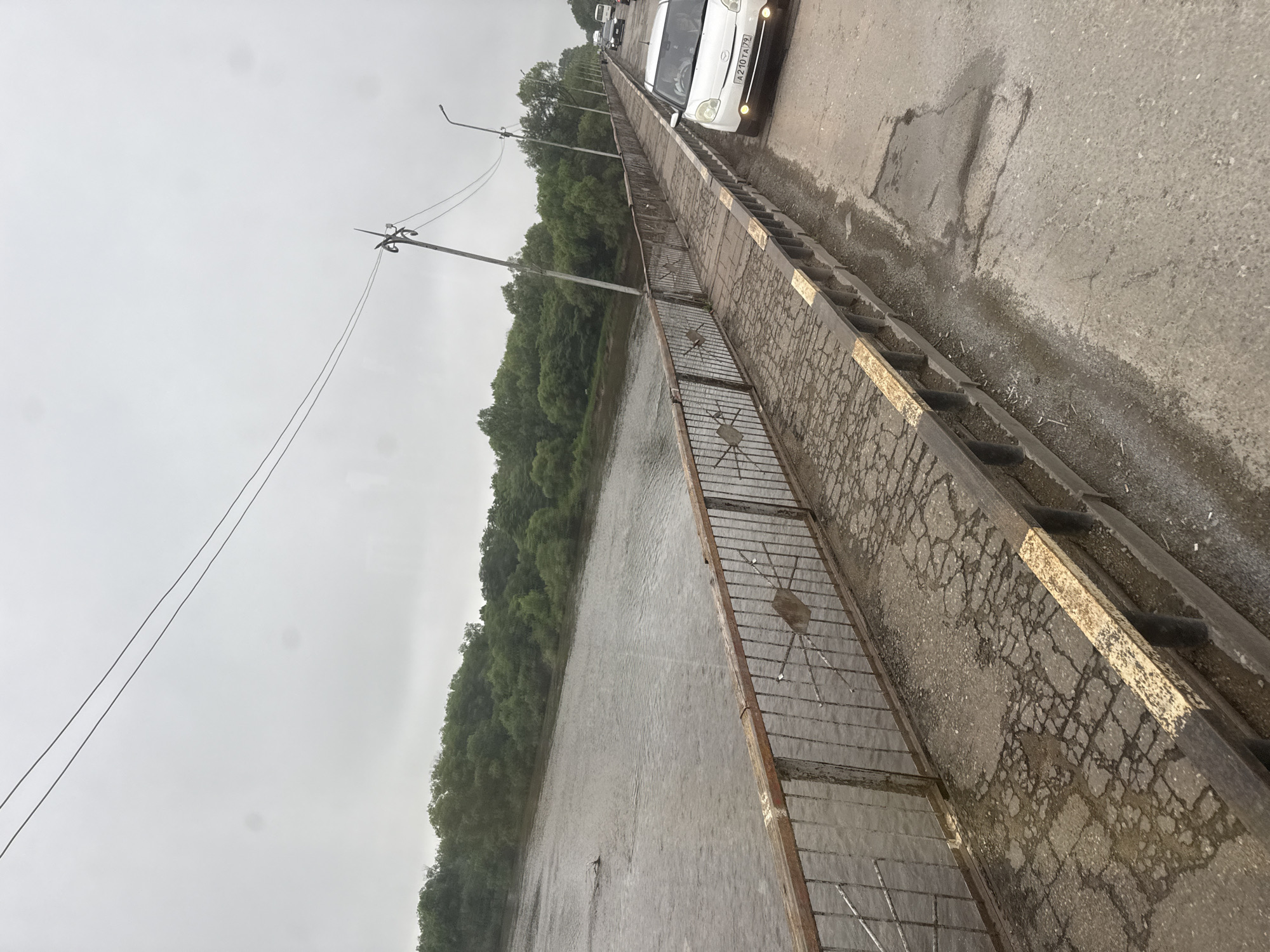 The image shows a bridge over a body of water, likely a river, under an overcast sky. The bridge has a metal railing with a wooden top and a cracked asphalt surface. There are trees lining the opposite bank of the water. On the bridge, there are several vehicles, including a white car with its headlights on and a license plate that reads "A 210 TA 79". Another car is visible further down the bridge, along with what appears to be a bus. Telephone poles with wires are present along the side of the bridge. The scene is consistent with the appearance of the Komsomolsky Bridge in Birobidzhan, Russia, which spans the Bira River and connects the city to the main highway. The weather appears damp, possibly indicating recent rain.