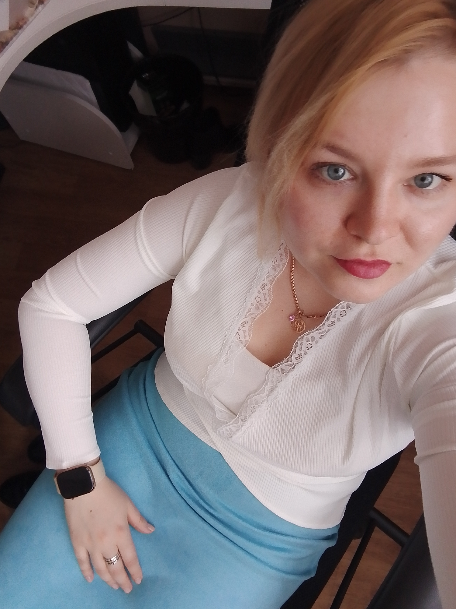 A blonde woman sits in a black chair, wearing a white, long-sleeved shirt with lace trim and a blue skirt. She wears a watch on her left wrist and a ring on her left hand. The background includes a wooden floor, a white shelf-like structure, and dark objects. The location is Izhevsk, Russia, based on visual elements characteristic of the region.