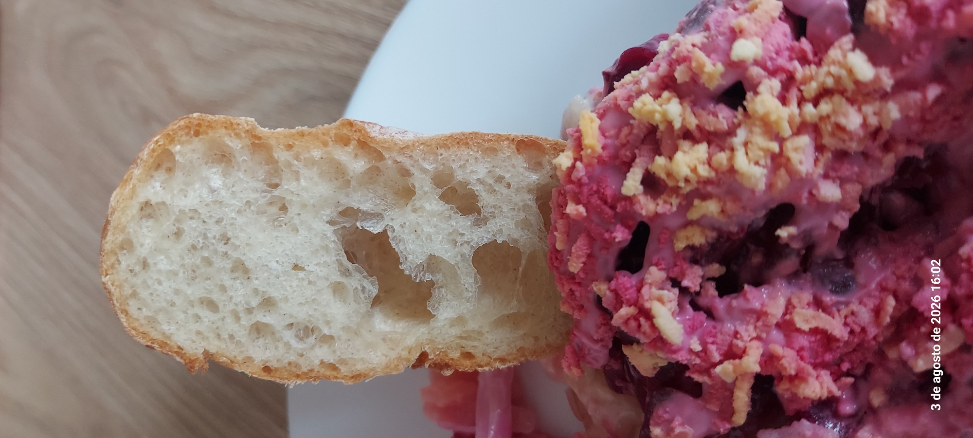 A close-up, eye-level, vertical, indoor shot captures a slice of crusty, white bread, likely from a baguette, partially obscuring a dish of what appears to be a beet-based salad. The bread, with its porous interior and golden-brown crust, is positioned in the upper half of the frame, tilted slightly. Below it, the salad is a vibrant mix of pink and dark red, topped with crumbled yellow bits, possibly hard-boiled egg or cheese. The texture of the salad is creamy and granular. In the lower left corner, a timestamp "3 de agosto de 2026 16:02" is visible, suggesting the image was taken on August 3, 2026, at 4:02 PM. The background consists of a neutral, light-colored surface, possibly a table. The scene suggests a casual meal setting. The location of Castelldefels, Spain, is not visually evident in the provided image.