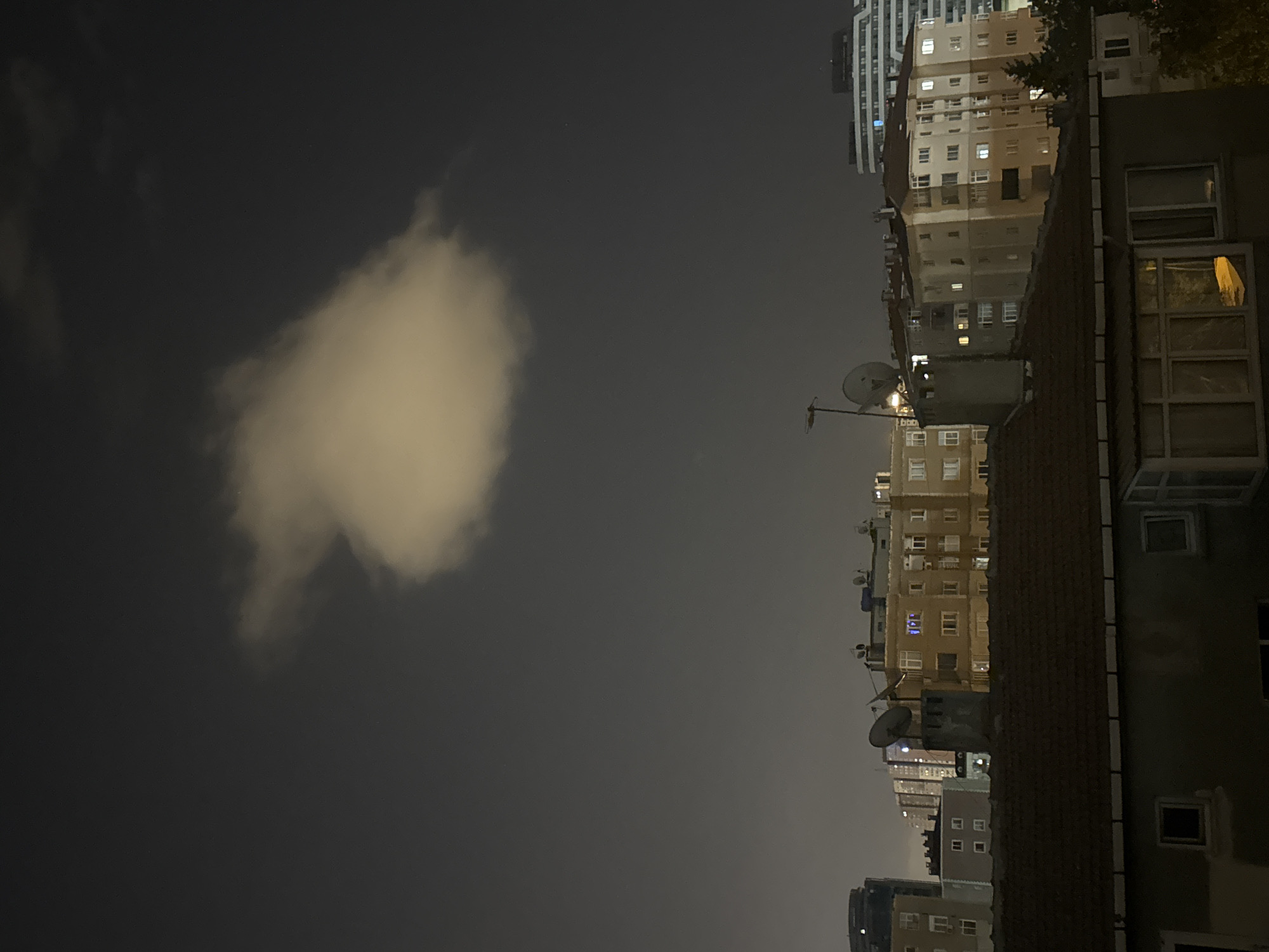 This is a nighttime photograph taken from a rooftop in Gungoren, Turkey. The sky is dark and overcast, with a single, illuminated cloud formation near the top center of the image. Below the cloud, a dense cluster of apartment buildings dominates the middle ground. These buildings vary in height and architectural style, with many windows illuminated by interior lights. Satellite dishes are visible on some of the rooftops. In the foreground, the roof of a lower building is prominent, featuring brown shingles and dark walls with visible windows. The overall impression is of a densely populated urban environment at night.