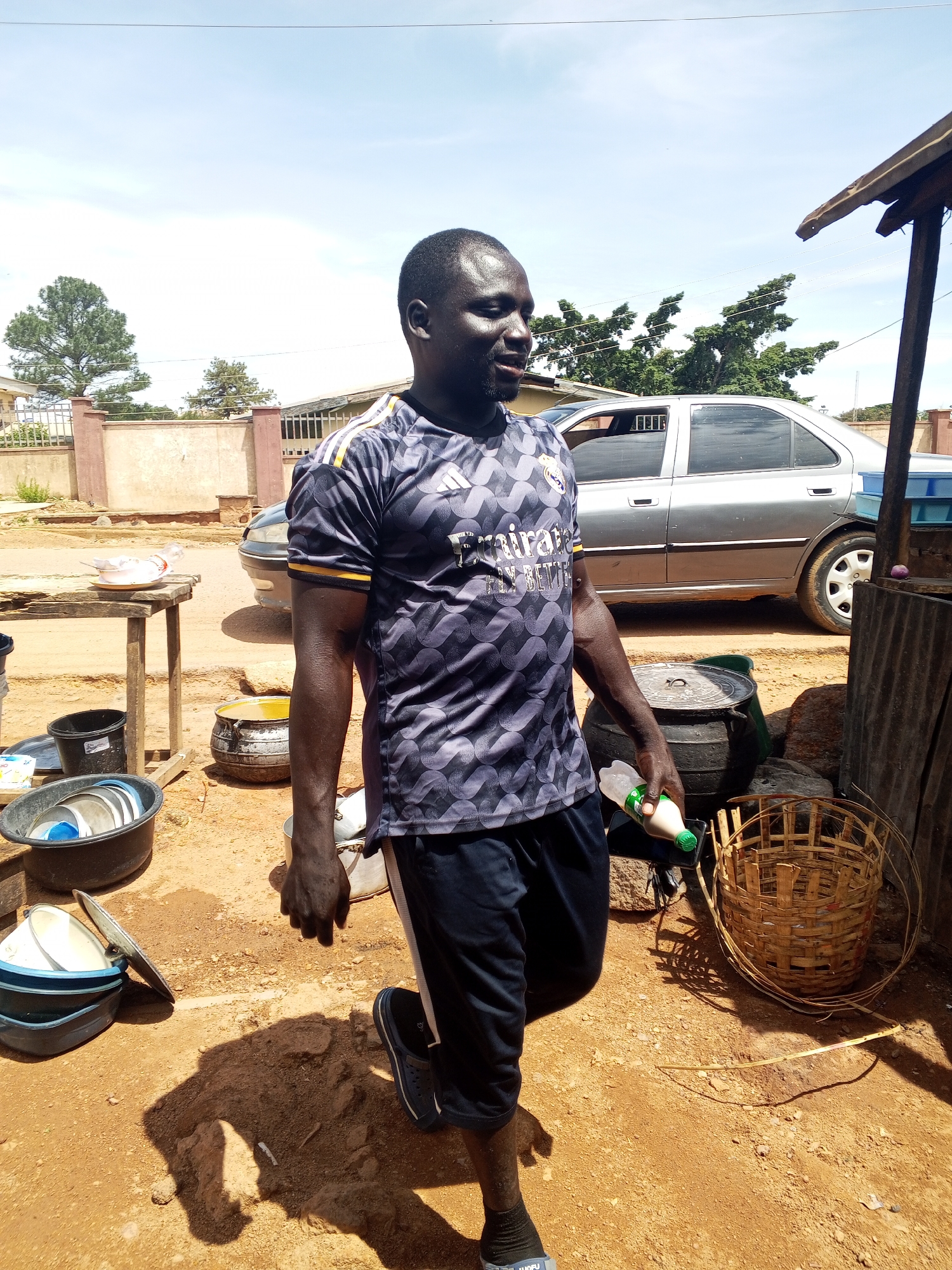 This outdoor scene, set in the city of Jos, Nigeria, features a dark-skinned adult male walking across unpaved reddish-brown dirt ground under a clear blue sky. He wears a grey and black patterned Real Madrid soccer jersey with "emirates FLY BETTER" text, dark shorts, and black sandals with socks. In his right hand, he holds a white plastic bottle with a green cap. To his left, a wooden table holds a white plate and a plastic bottle. On the ground near the table are black plastic basins containing dishes and a black pot with a lid, partially filled with a yellow liquid. In the background, a silver sedan is parked, and residential buildings with fences are visible among green trees. To the far right, a woven bamboo basket and a corrugated metal structure are present. The sunlight casts distinct shadows on the ground.