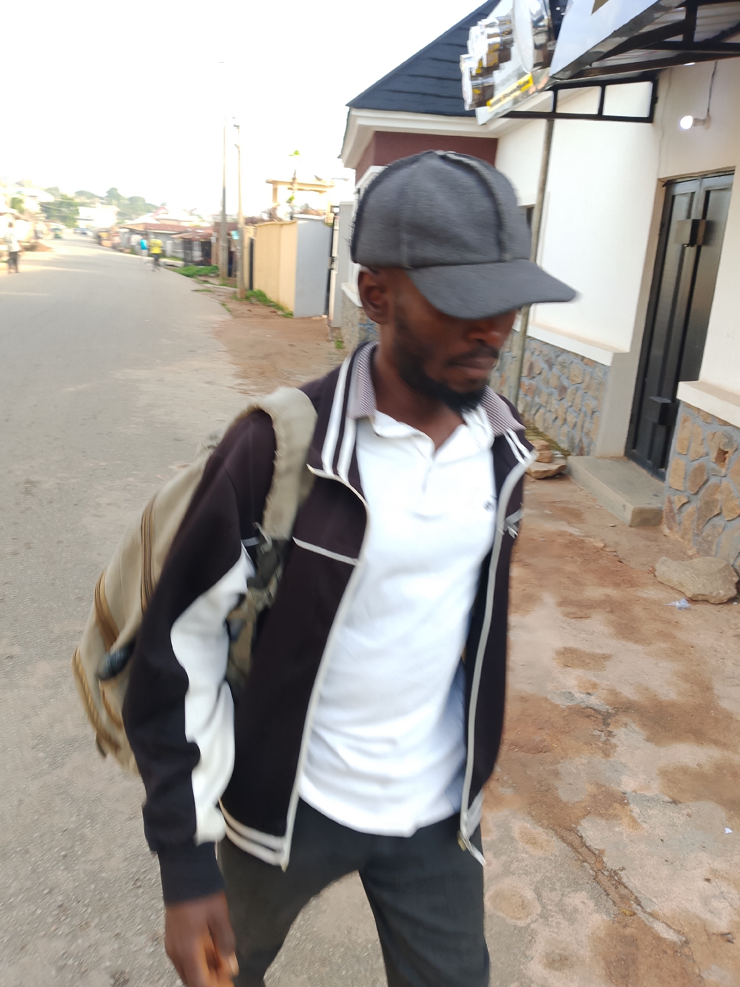 A man wearing a grey baseball cap, a white polo shirt, a black and white jacket, and dark pants, walks down a paved street in Jos, Nigeria. He is carrying a light brown and beige backpack over his left shoulder. The street is lined with buildings, some with stone accents and others with a cream-colored facade. In the background, other buildings and trees are visible, with a bright, overcast sky.