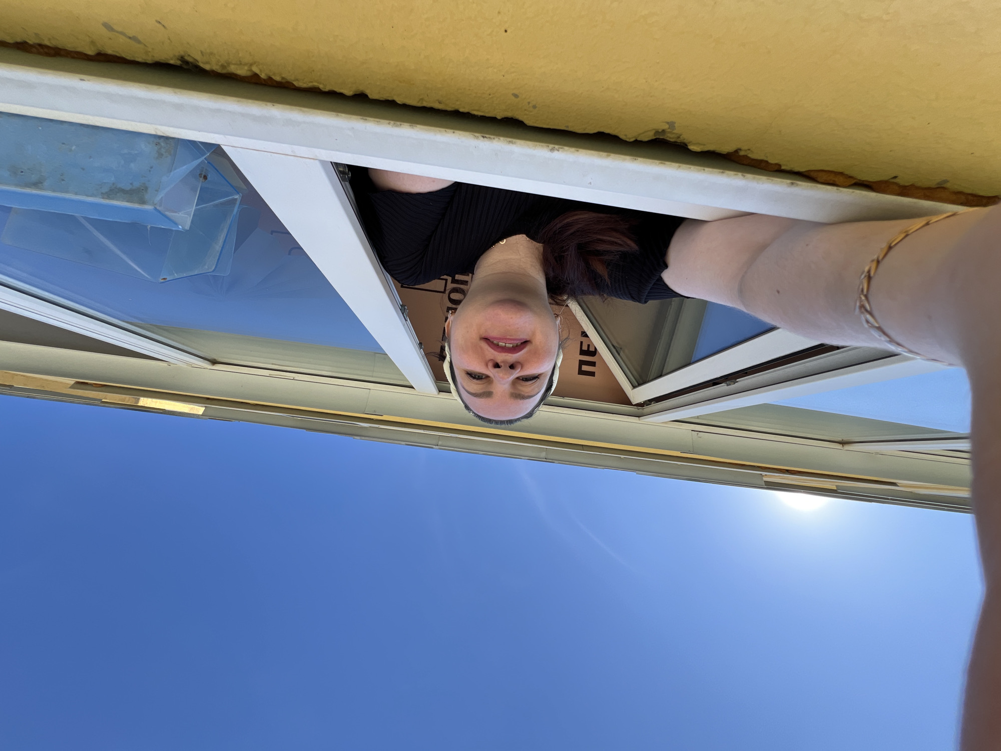 A woman is partially visible looking out of a window. The background shows a clear blue sky and the exterior of a building. The woman has fair skin and dark hair. She is wearing a black top. A gold bracelet is visible on her arm. The window is white framed.
I am unable to determine the location in the city of Noviy, Russia as it is not possible to extract specific location information from this image.