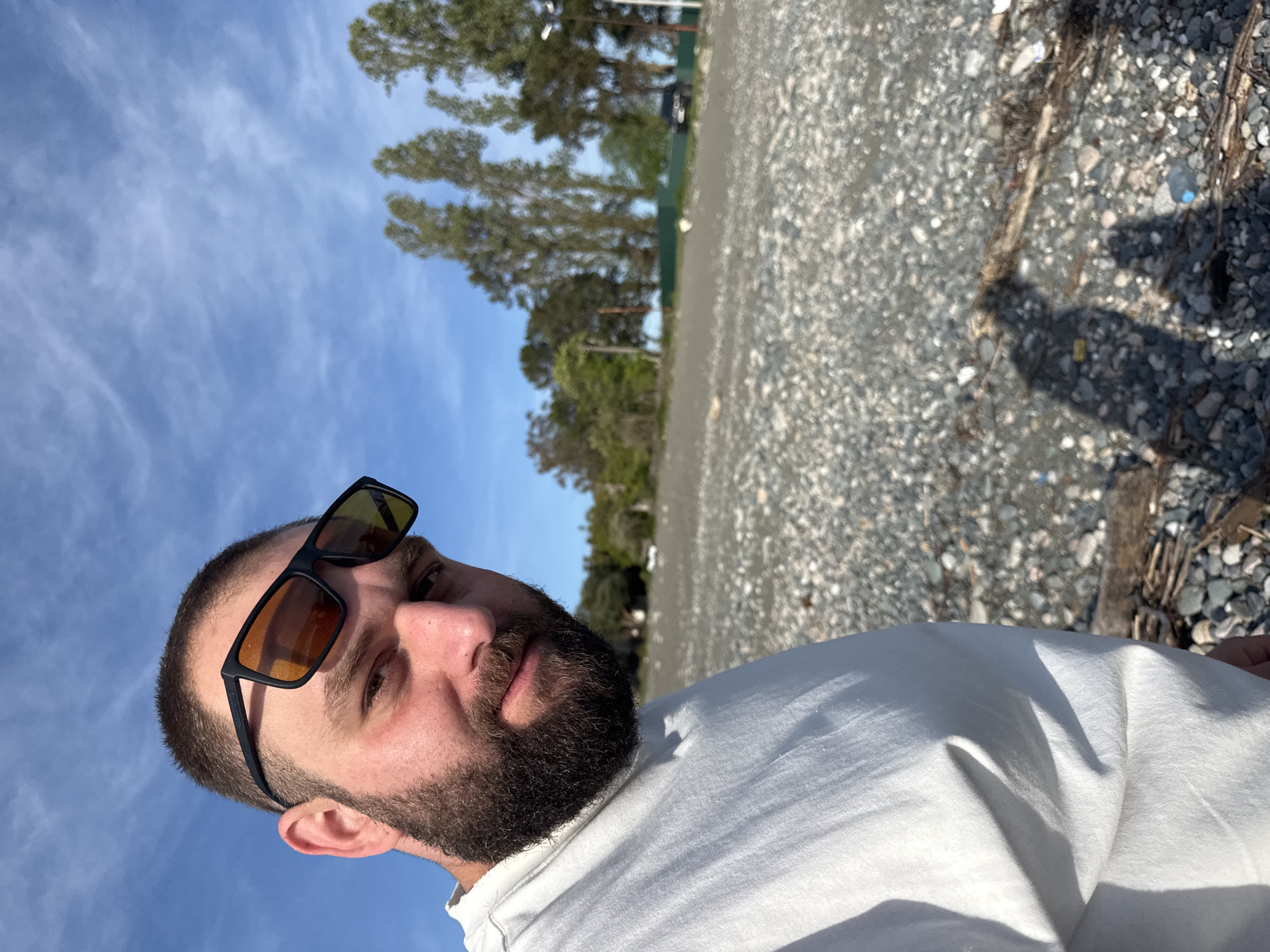 A man with a beard and sunglasses is posing for a selfie in Yuada-Gulrypsh, Abkhazia. The backdrop is a gravelly beach with scattered driftwood and trees. The sky is blue with scattered clouds. The man is wearing a white shirt.