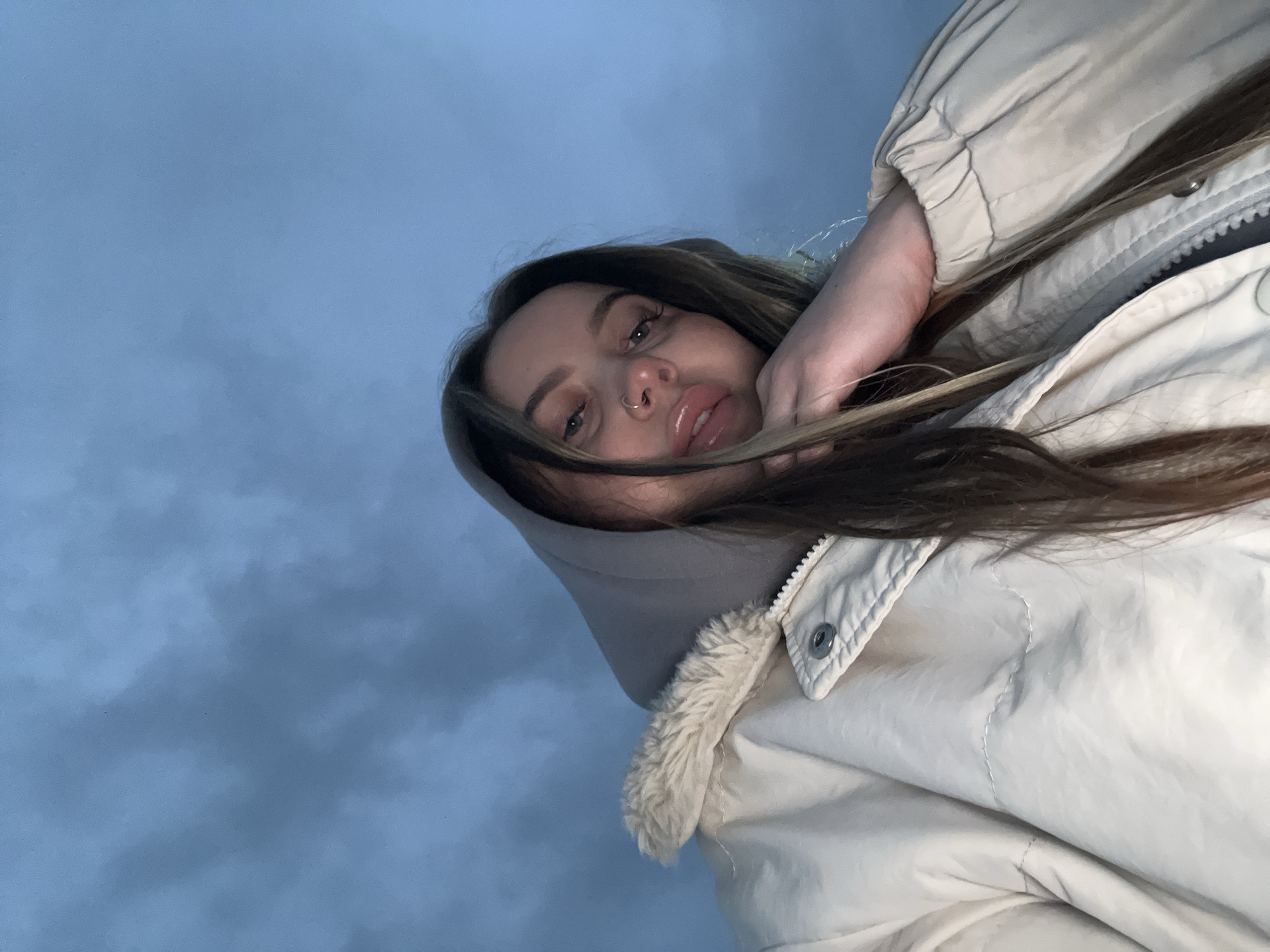 A young woman with brown hair and fair skin is seen from a low-angle perspective against a backdrop of a cloudy sky. She has a nose ring and is wearing a light-colored jacket with a furry collar and a hood. Her right hand is partially obscuring her face.
I am unable to determine the location in the city of Novopokrovskaya, Russia, due to a lack of discernible landmarks or contextual information.