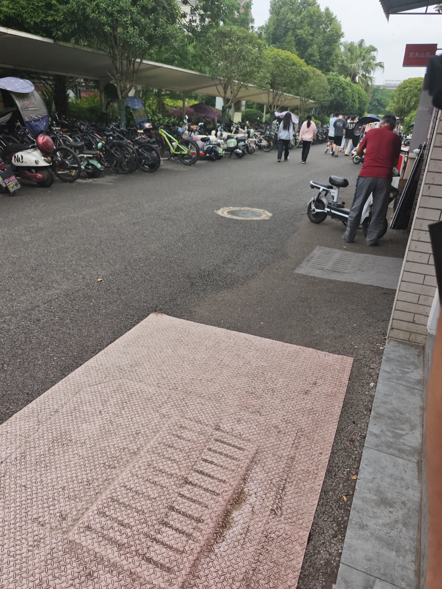 The image depicts a scene in the Chongqing High-tech Zone, China, featuring a parking area for electric scooters and bicycles. In the foreground, a large, textured metal plate lies on the asphalt. Further back, rows of parked electric scooters and bicycles are visible, some covered by umbrellas. A few individuals are walking in the background, some carrying umbrellas. One person in a red shirt and grey pants is standing next to an electric scooter. Trees and a covered walkway line the parking area. A red sign with Chinese characters is mounted on a building in the upper right corner. The overall environment appears to be an outdoor public space in a city setting.