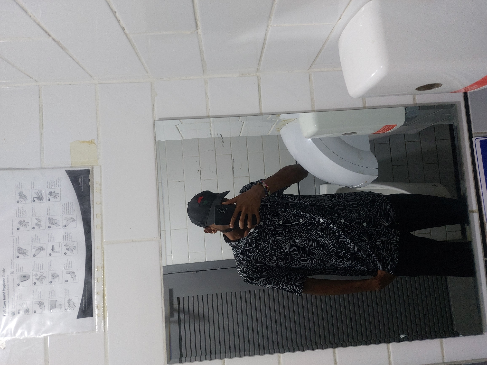 A person is taking a selfie in a restroom mirror. The individual is wearing a black baseball cap, a black shirt with white swirling patterns, and black pants. In the mirror, a white sink, paper towel dispenser, and a grey slatted barrier are visible. To the left of the mirror, a hand hygiene guide is affixed to the wall. The restroom is tiled with white square tiles. The location is Dar es Salaam, Tanzania.