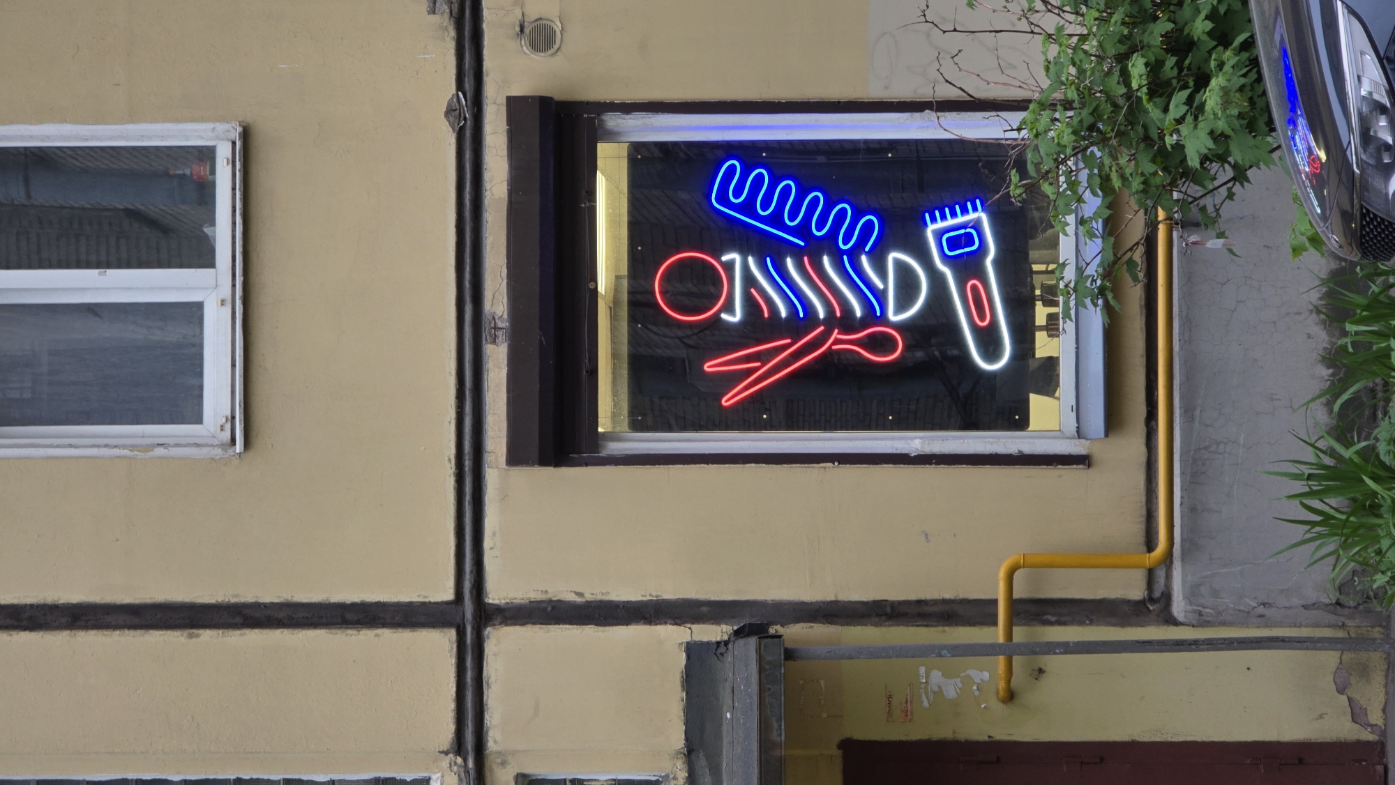 The image depicts the exterior of a building with a barber shop sign in a window. The building is painted in a pale yellow color with dark horizontal and vertical lines outlining rectangular sections. To the left of the barber shop window is a smaller, white-framed window. Below the barber shop window is a dark gray and beige door or hatch. A bright yellow gas pipe runs vertically along the right side of the barber shop window and then horizontally.
The barber shop sign is a neon display featuring a blue comb, red scissors, and a white razor. The window reflects the interior of the shop.
On the right side of the image, green foliage partially obscures the view of the building and window. Part of a silver vehicle is visible near the top right corner. The location is potentially in Saint Petersburg, Russia, given the building's architectural style.